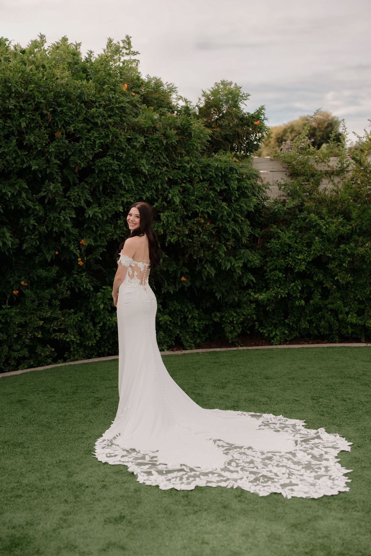 Elegant white wedding dress with lace train photographed on green lawn with hedge backdrop.