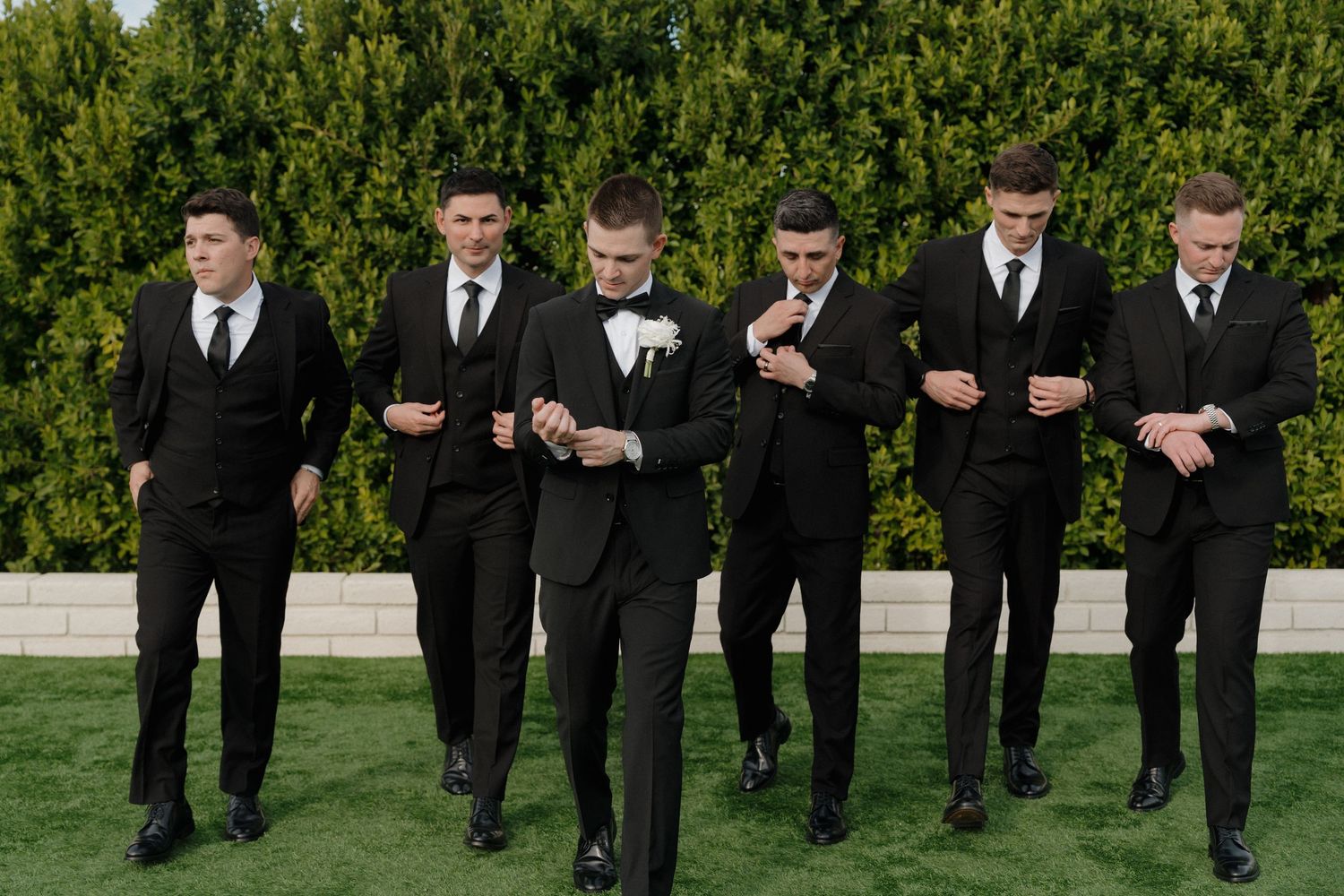 A group of groomsmen in black suits stand together on a lawn in front of a green hedge wall.