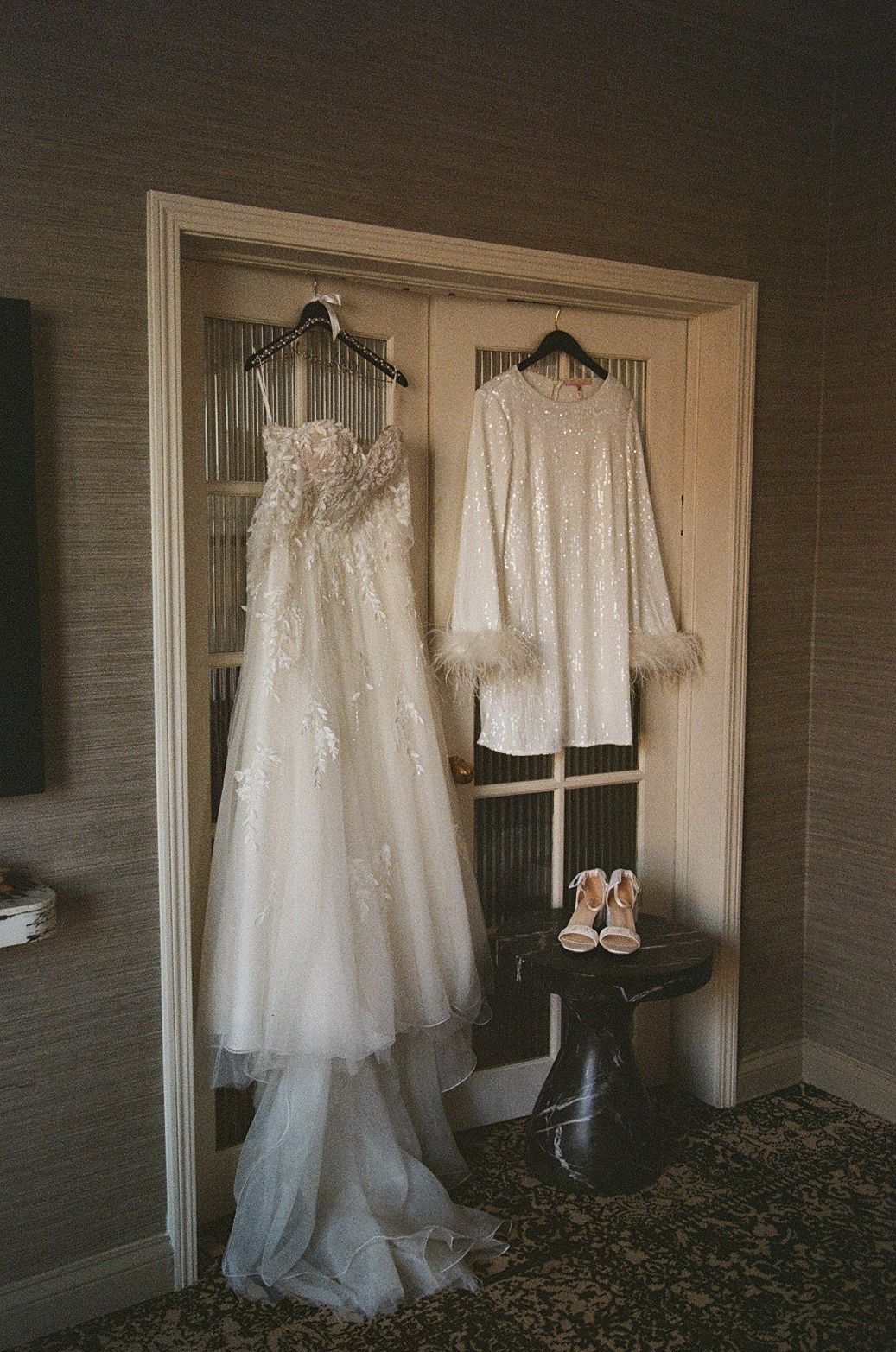 Bridal gown and sequined jacket with feather cuffs hanging in doorway, with white heels on stool below.