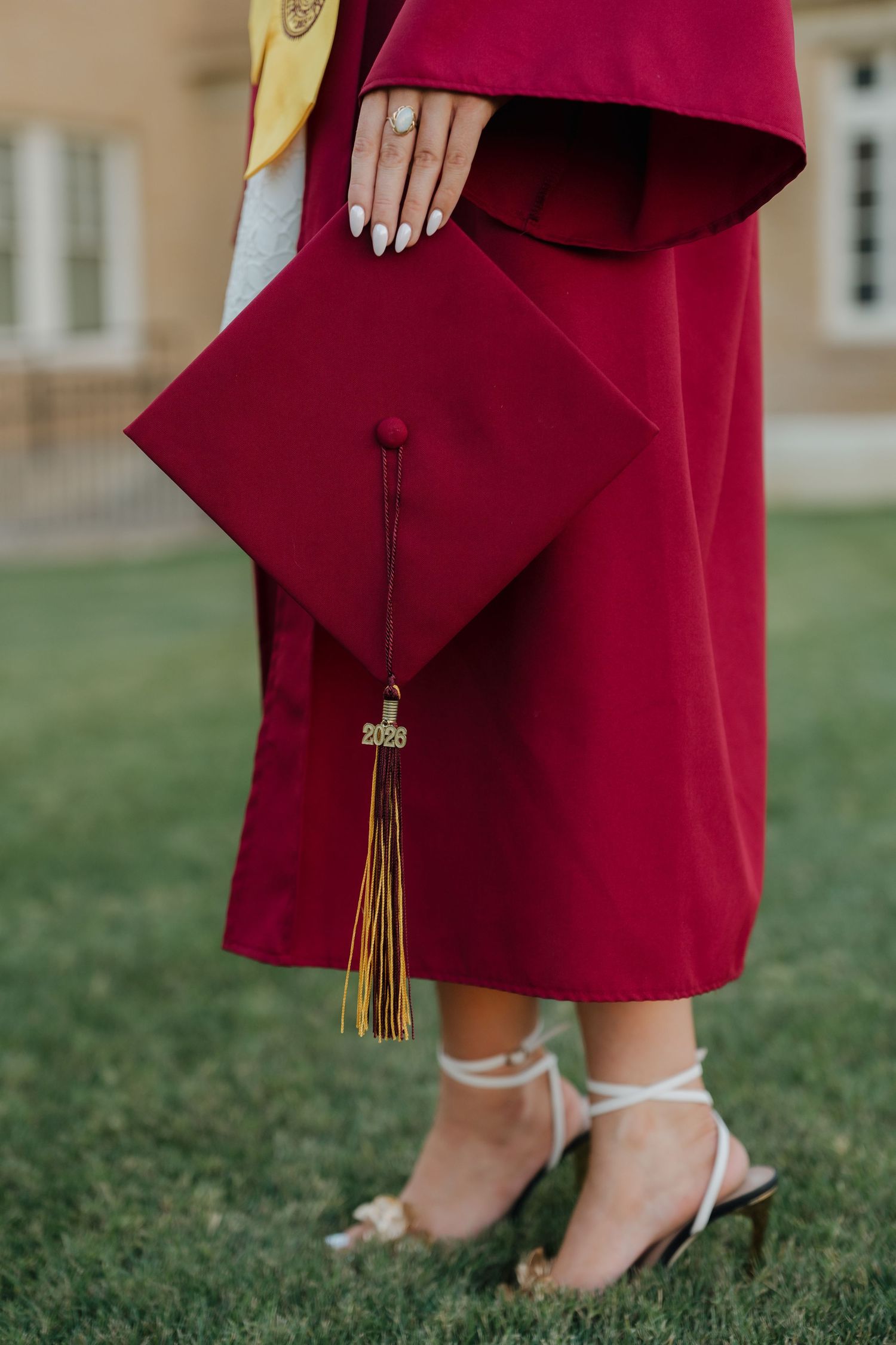Graduate in crimson cap and gown holding mortarboard with gold tassel and 2023 charm, wearing white heels.