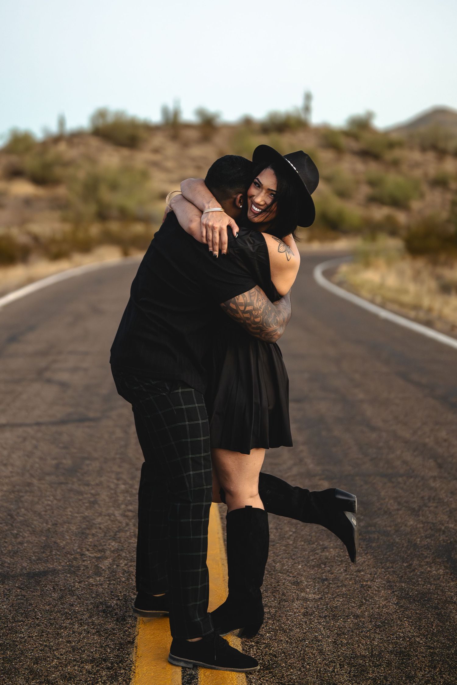 A romantic embrace between a couple standing on an empty desert road at sunset.