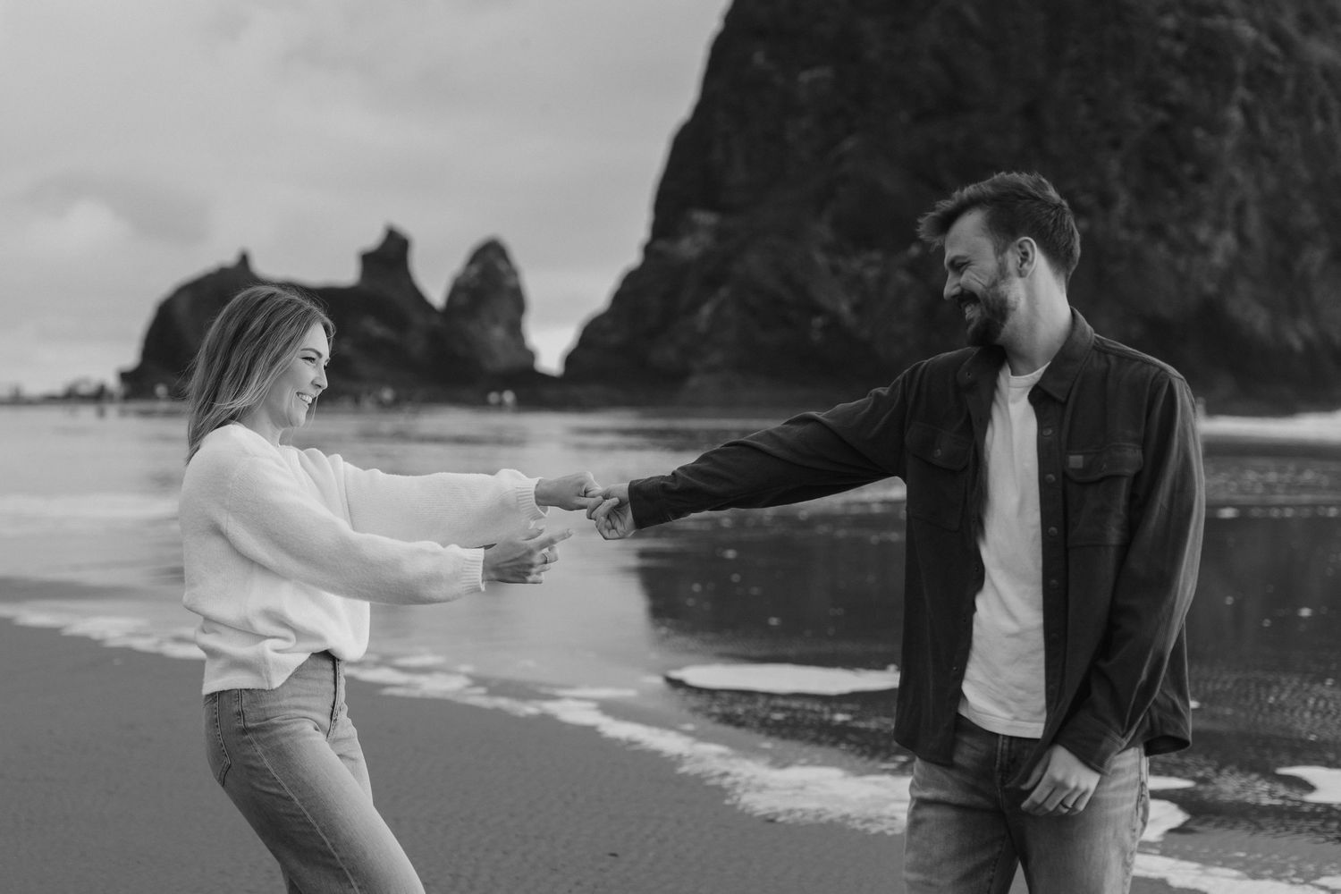 Black and white photo of a couple holding hands and laughing on a rocky beach with sea stacks in the background.
