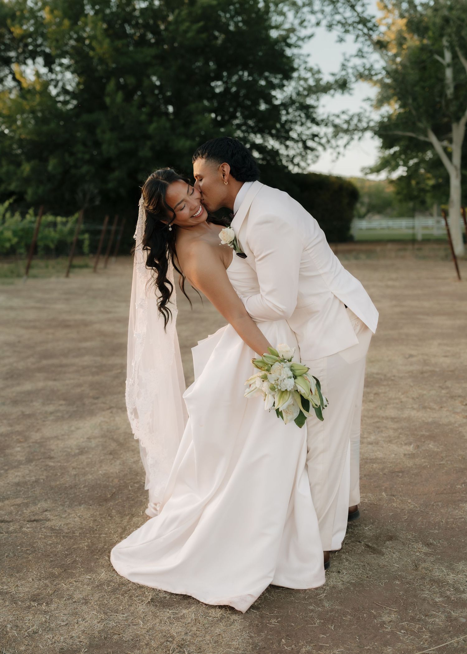 A groom in a cream suit kisses his bride in a white gown and veil while she holds a green and white bouquet.