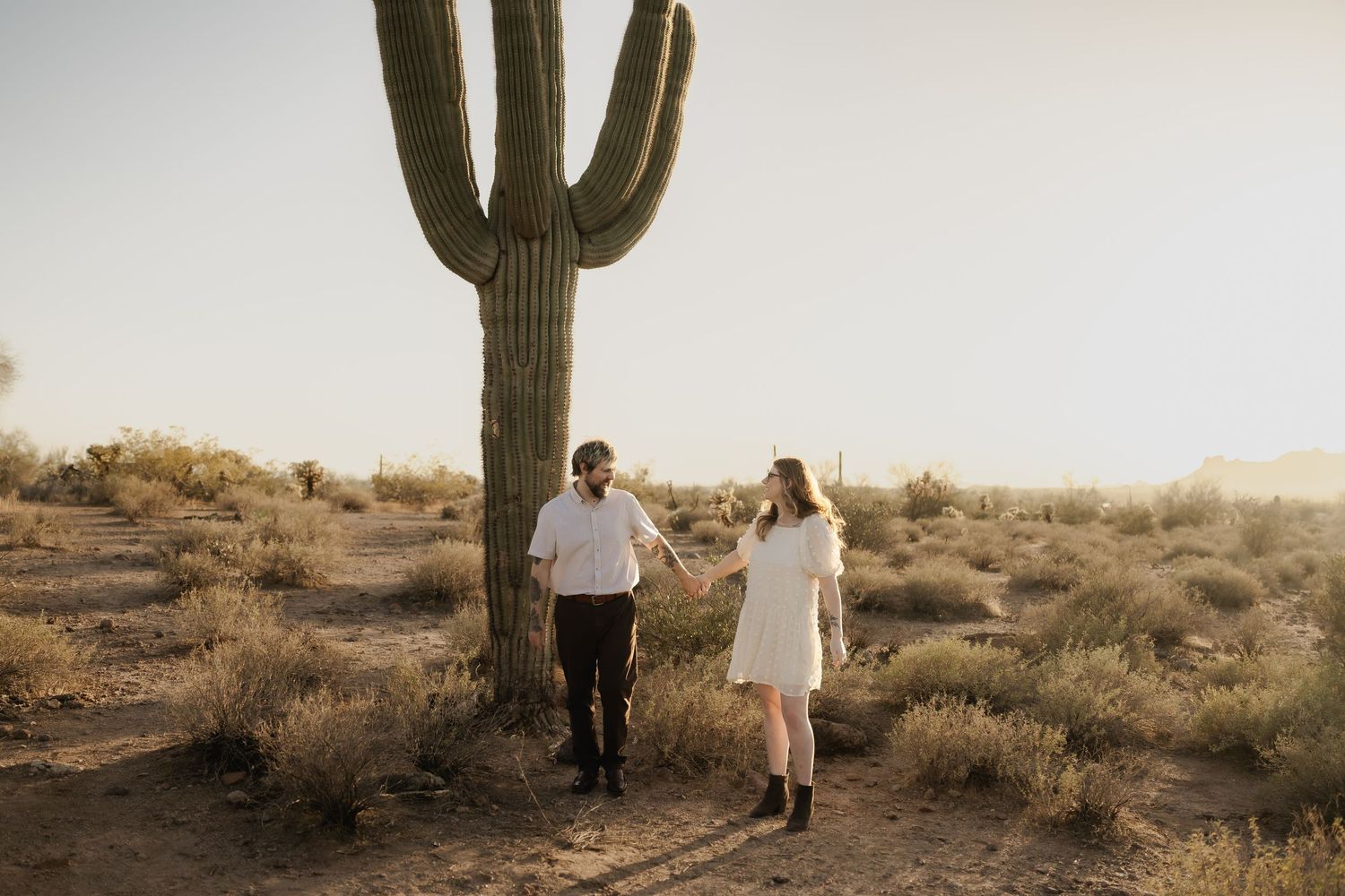 Desert engagement photo session with couple walking among tall saguaro cacti at sunset in the Arizona desert.