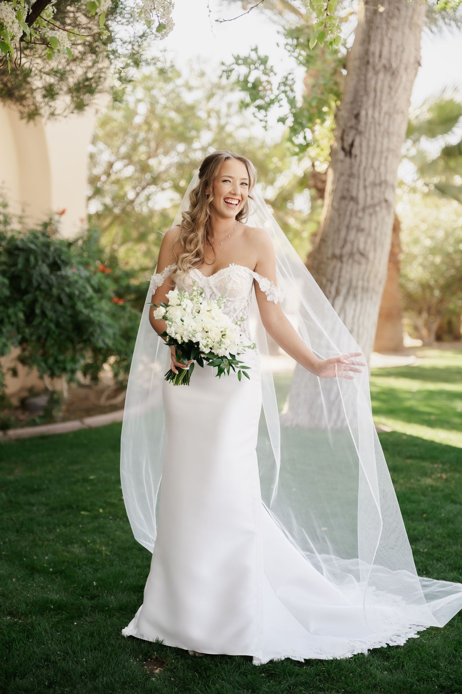 Elegant bride in a flowing white gown poses with her bouquet on manicured lawn.