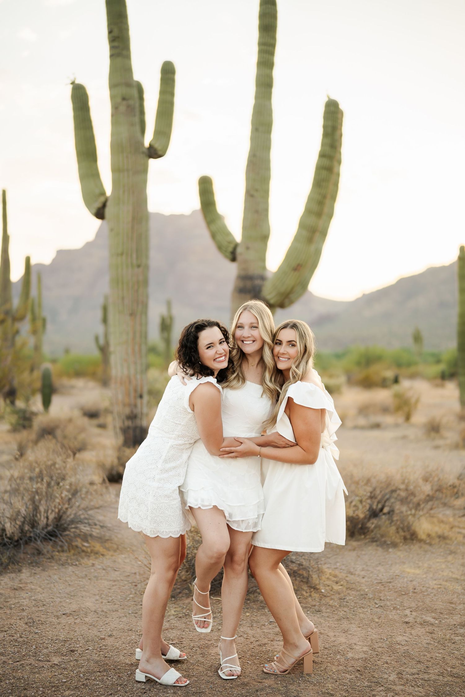 Friends in white dresses huddle together laughing in a desert setting with mountains in background.