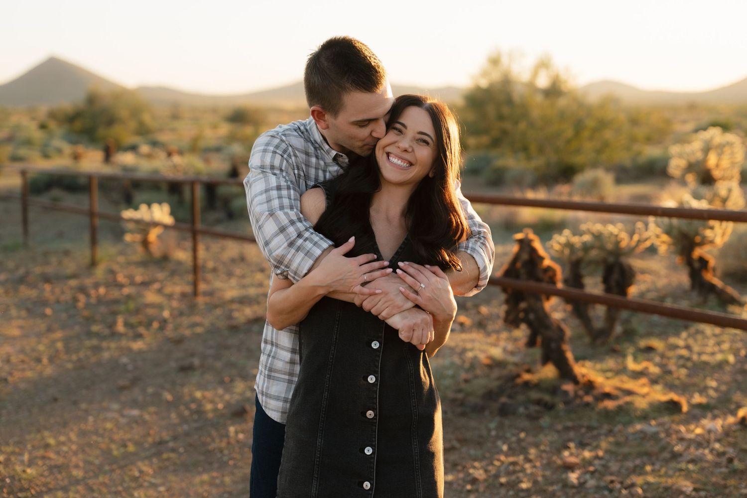 Couple embracing and laughing at golden hour in desert landscape with cacti and mountains in background.