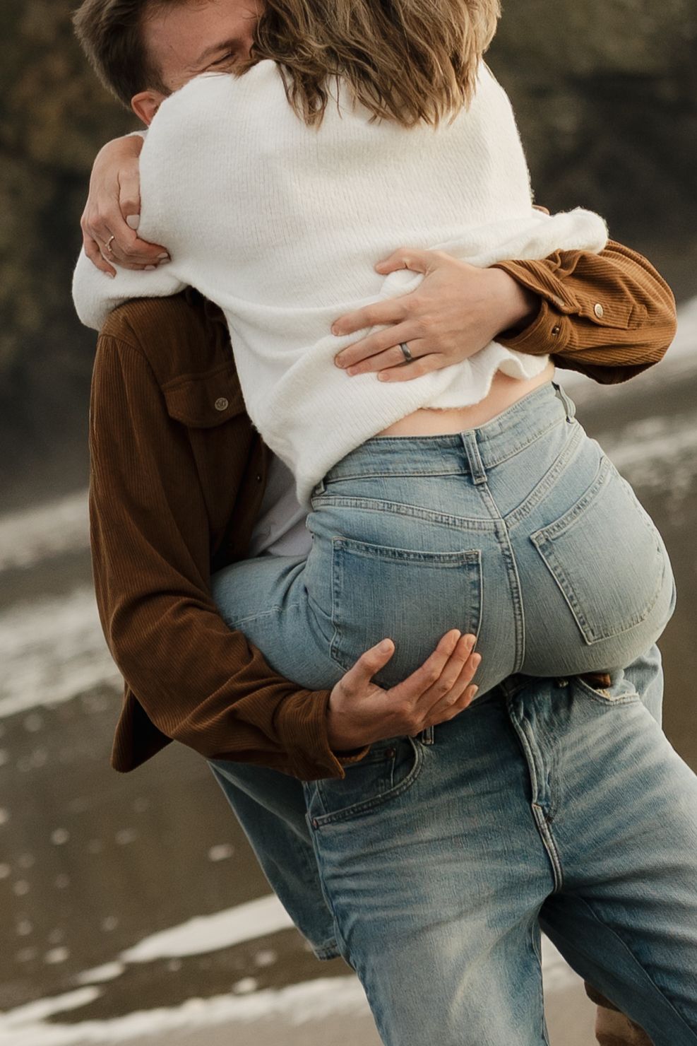 Couple embracing on a beach, woman in white sweater and jeans, man in brown jacket holding her close.
