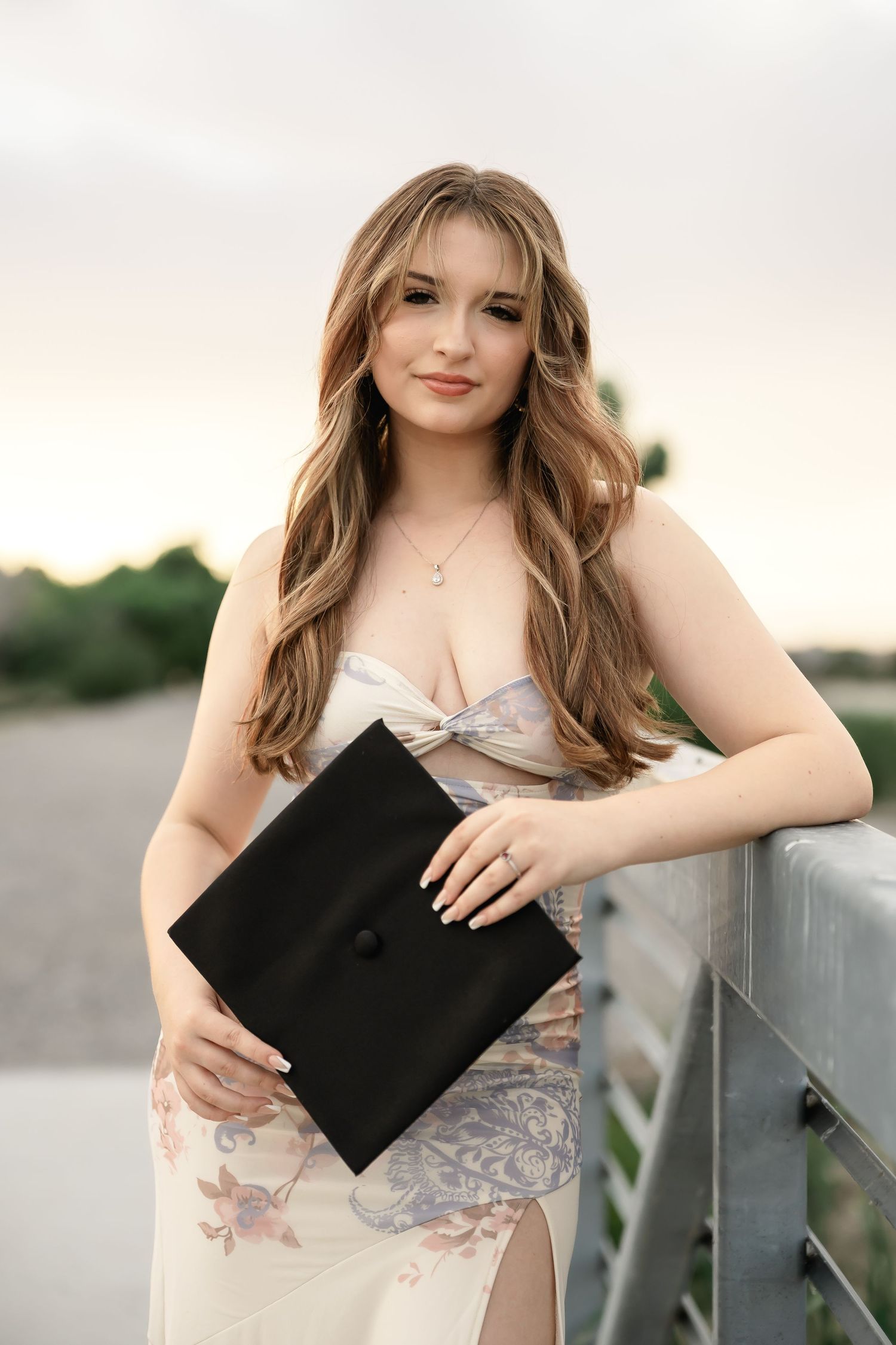 Someone in a floral dress poses with a black graduation cap against a soft cloudy background.