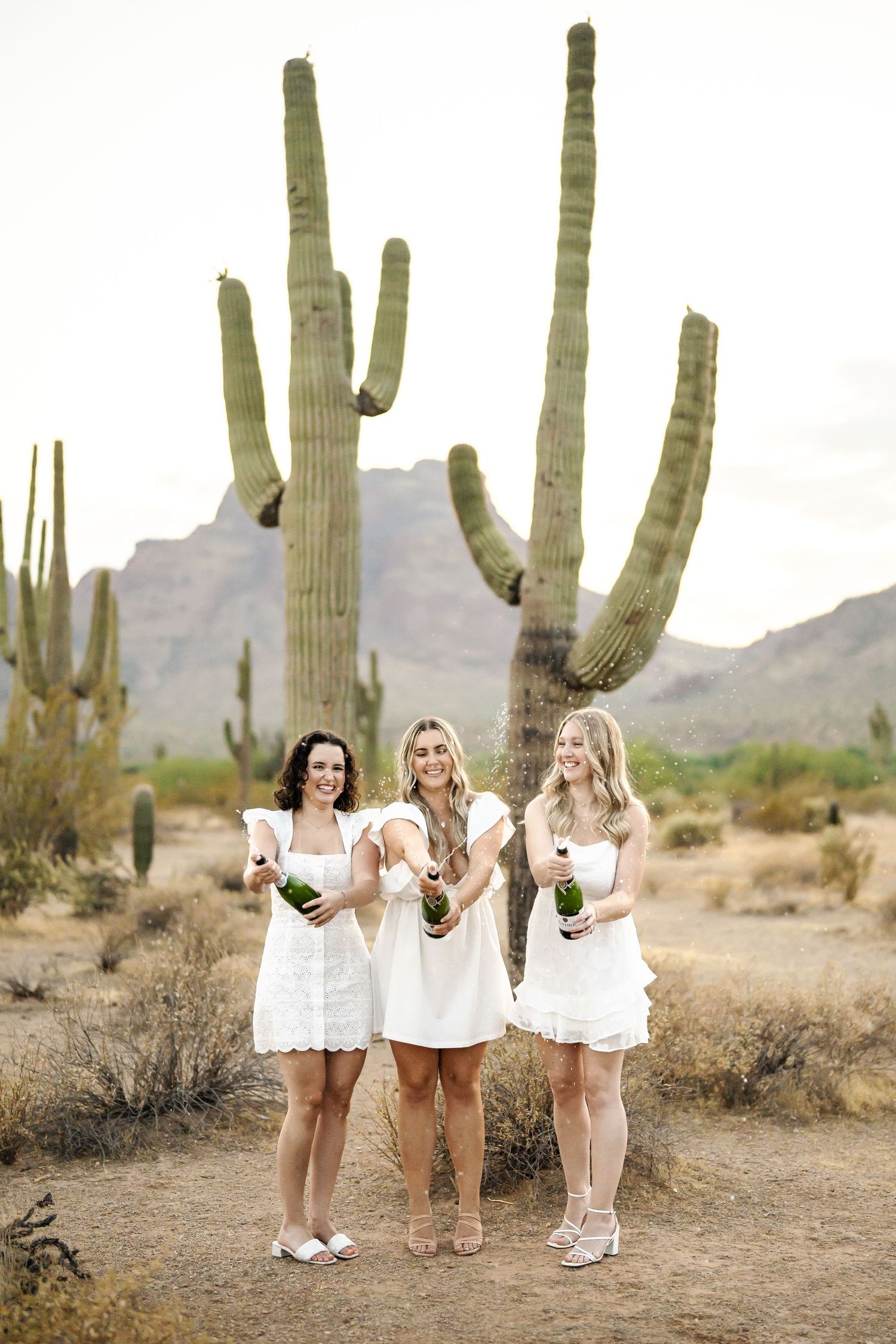 Friends in white dresses pose playfully with wine glasses in a desert landscape with saguaro cacti.