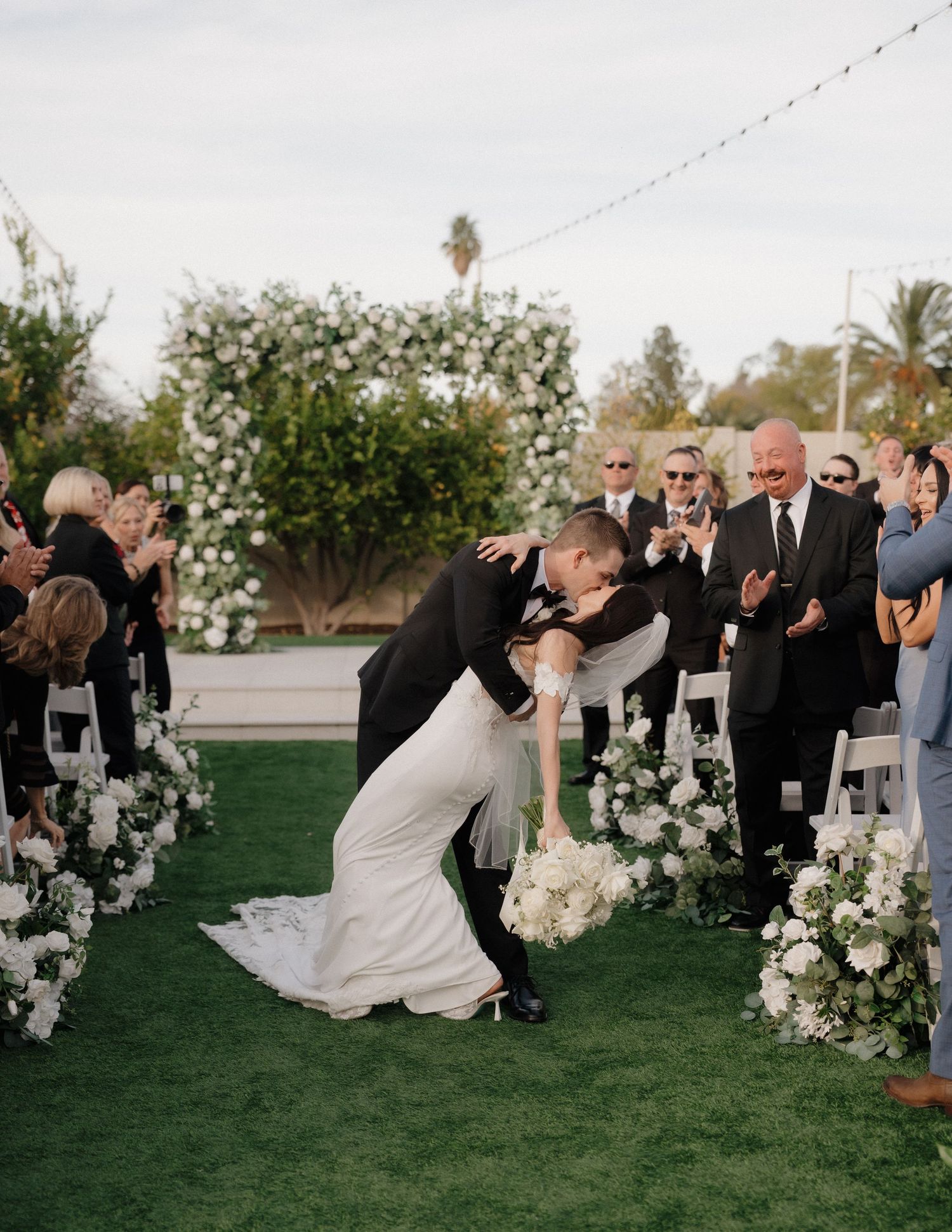 Wedding party performs a dramatic dip pose on a green lawn with white floral decorations at sunset.