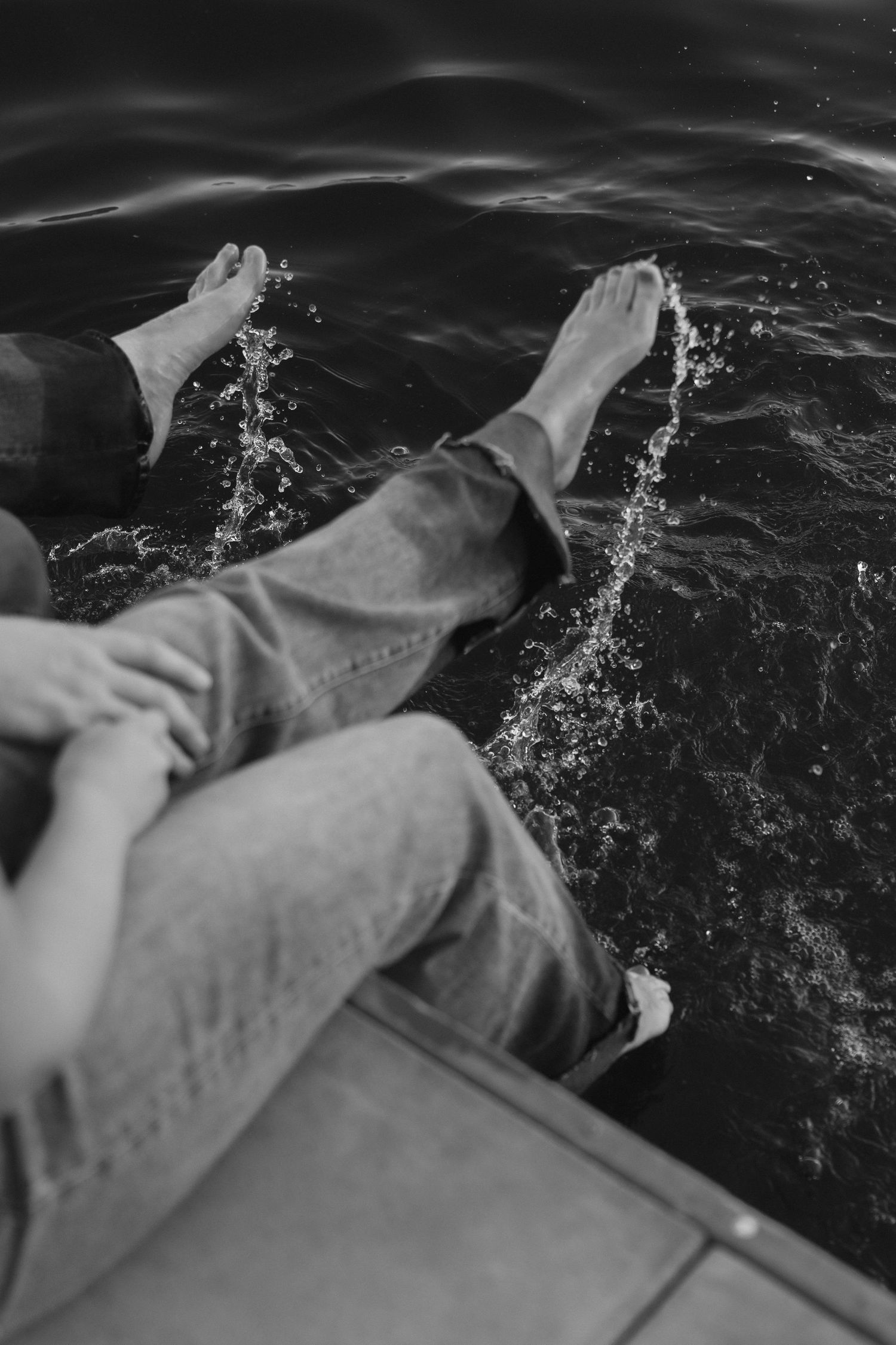 Black and white photo of a couple sitting on a dock, dangling bare feet over dark rippling water.