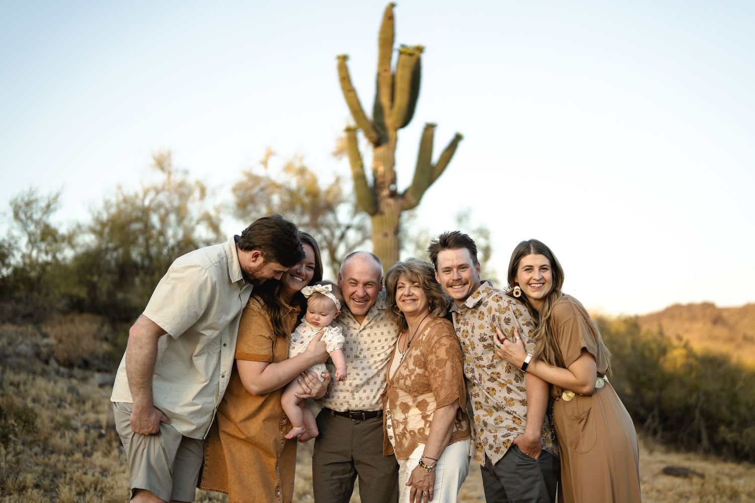 A family group in earth-toned clothing poses together at sunset in the desert with saguaro cacti in the background.