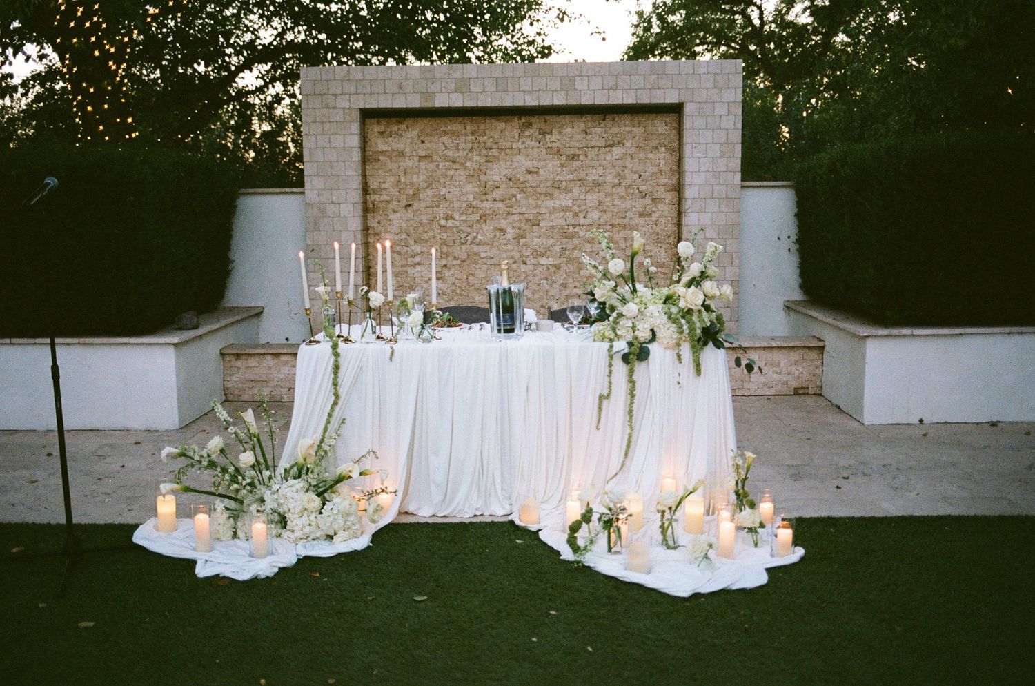 Elegant outdoor wedding sweetheart table with white draping, candles, and floral arrangements against a brick wall backdrop.