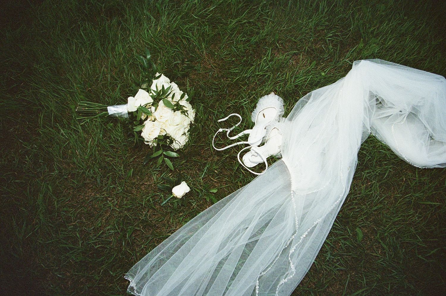 Bridal veil, white flower bouquet, and wedding shoes laid on lush green grass for a wedding flat lay.