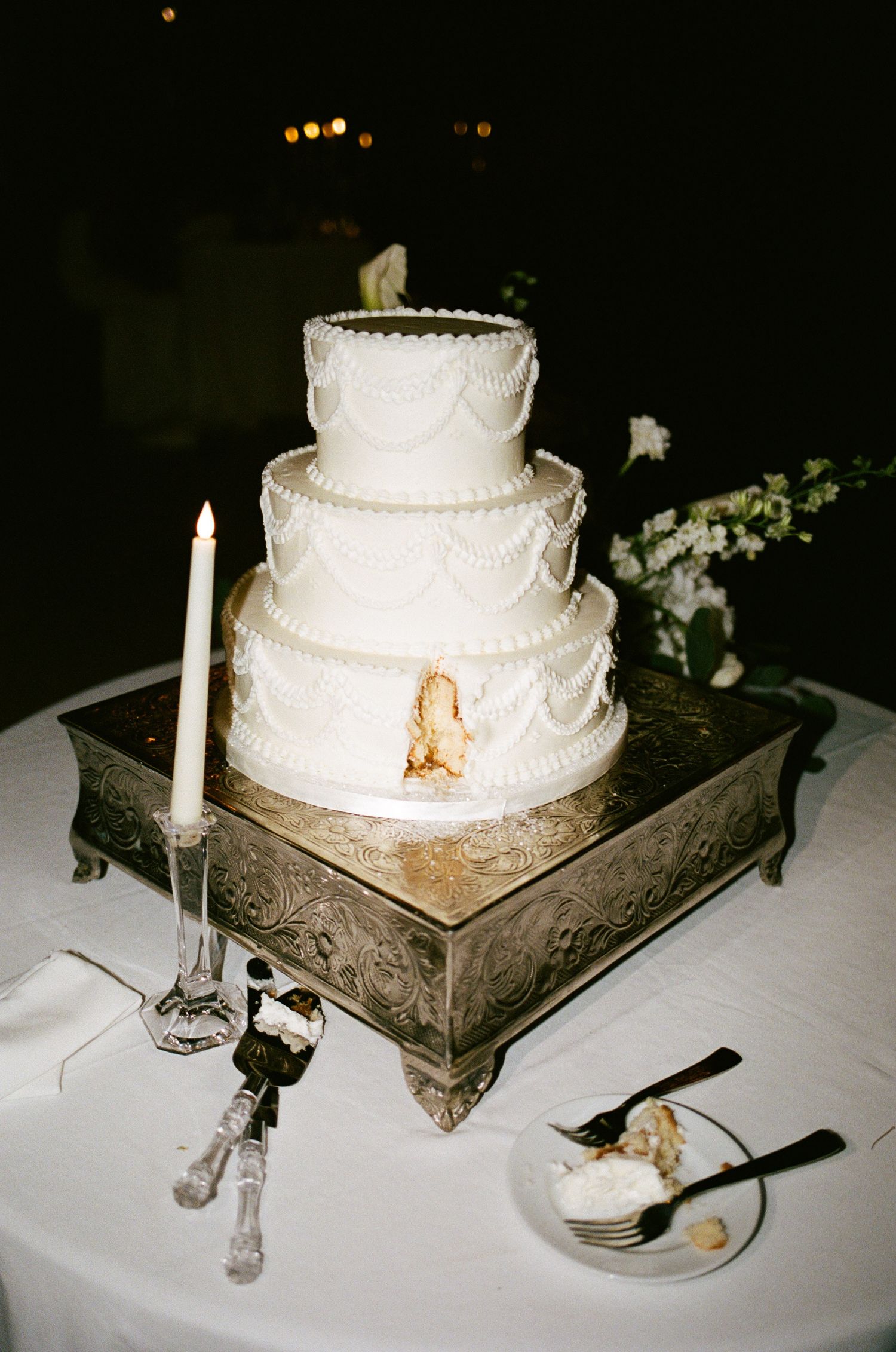 Elegant four-tier white wedding cake with draped frosting details on a silver stand, flanked by candles at a reception.