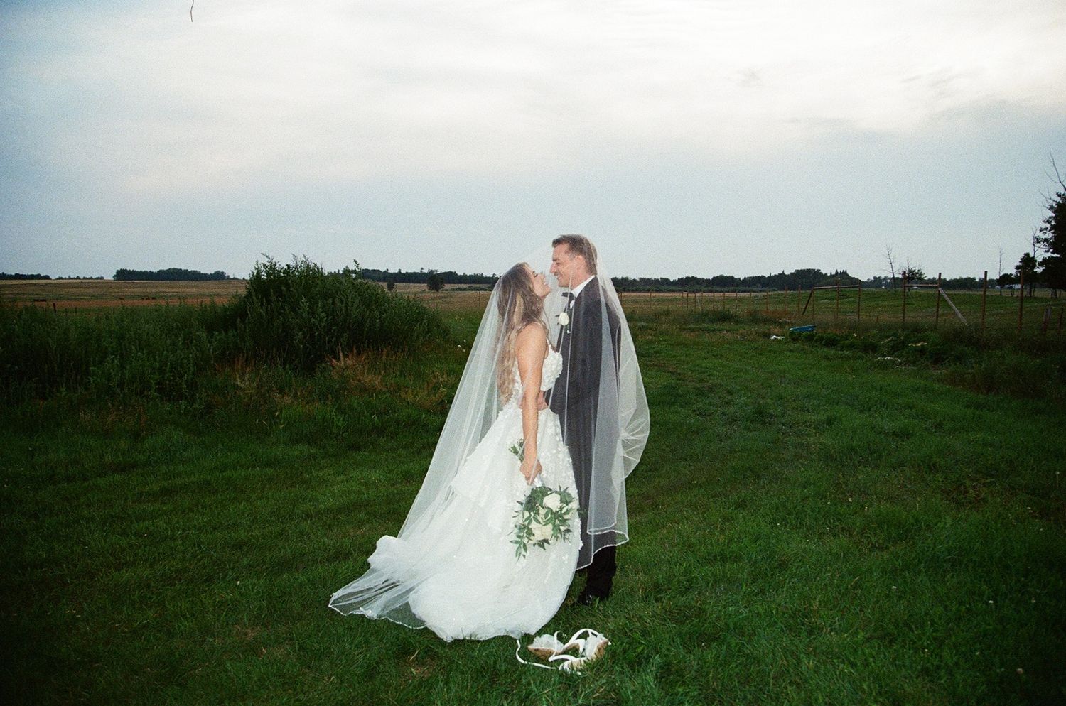 Bride and groom sharing a romantic kiss in a green field, bridal veil flowing in the wind on their wedding day.