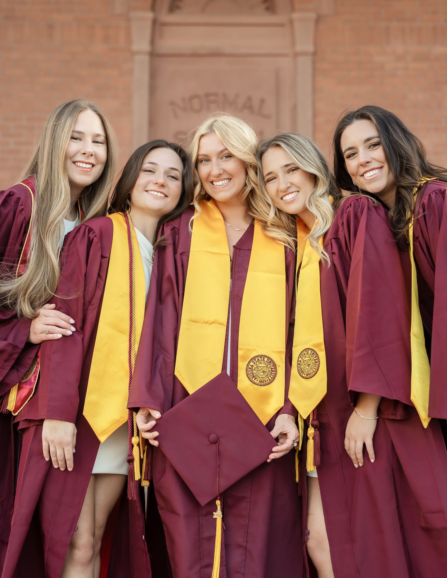 Row of graduates wearing maroon caps and gowns with gold stoles stand together for group photo.