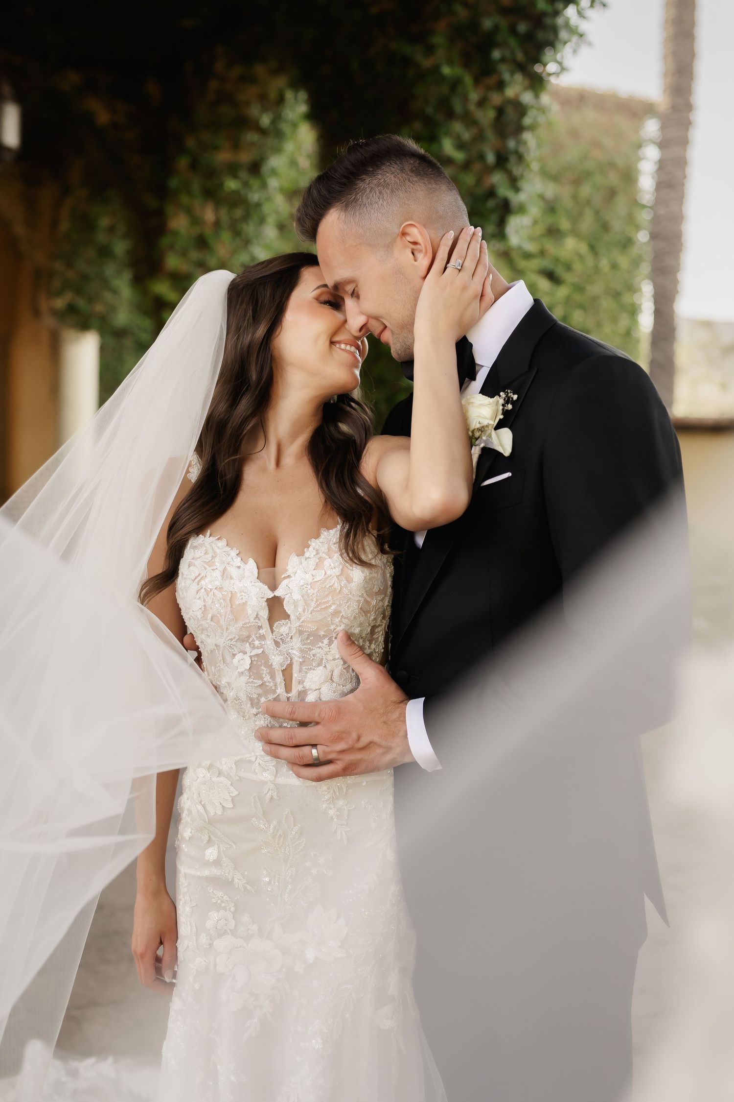 Bride in a lace wedding gown and veil embraces her groom in an intimate romantic moment outdoors.