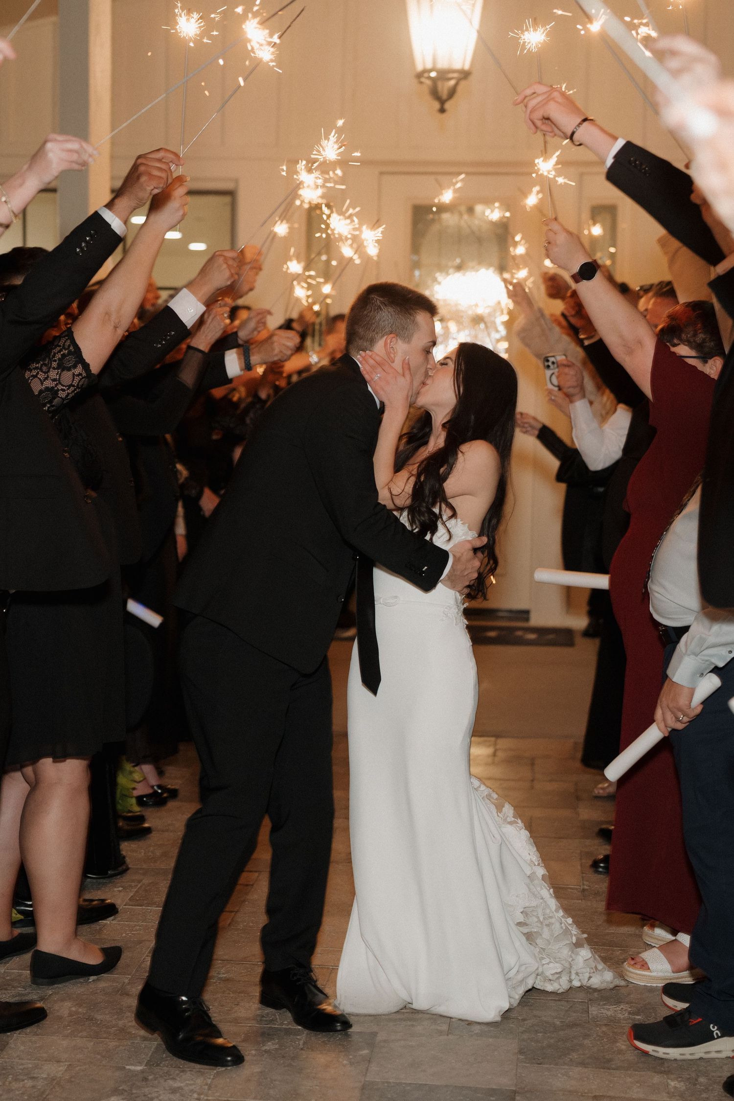 Wedding party guests wave sparklers in celebration during a nighttime reception at an elegant indoor venue.