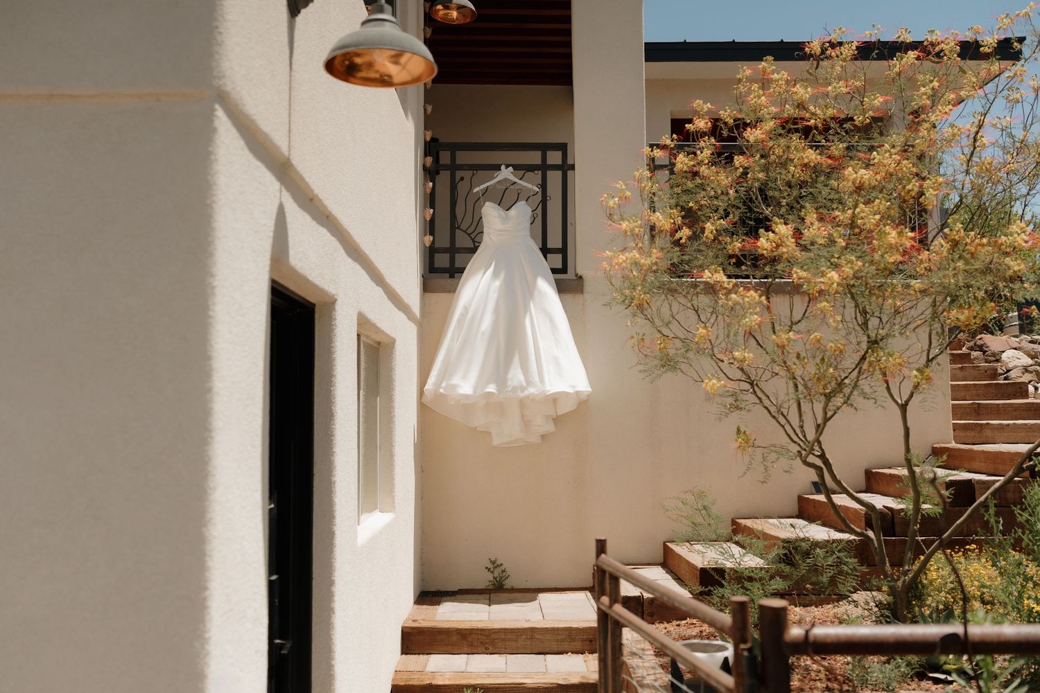 A white wedding dress hanging outside a rustic venue with stairs, greenery, and warm afternoon light.