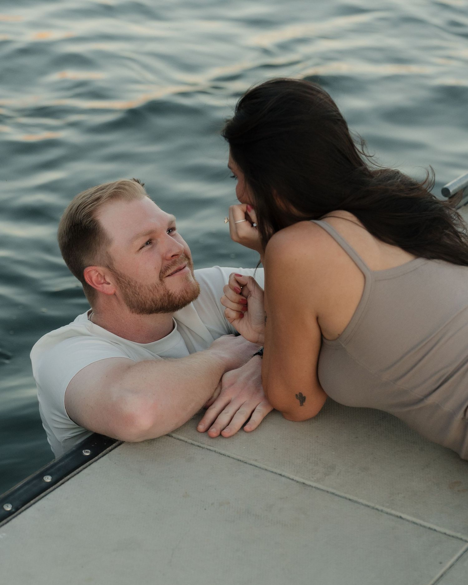 A couple shares a romantic moment by the water, with a man in the water leaning on a dock and a woman sitting beside him.