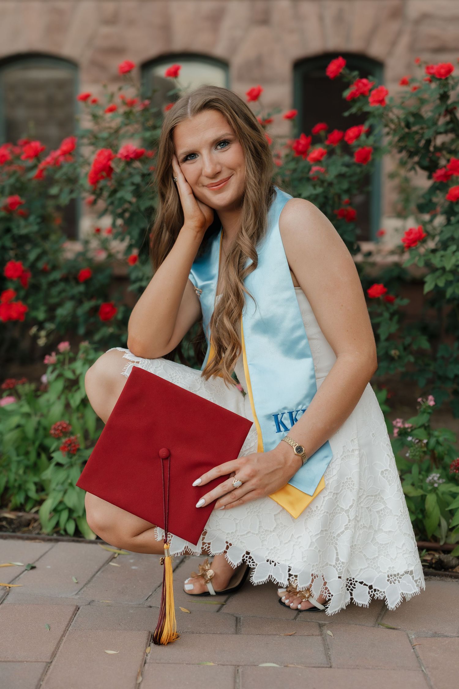 Young woman in white lace dress poses with red graduation cap amid blooming roses for senior photos.