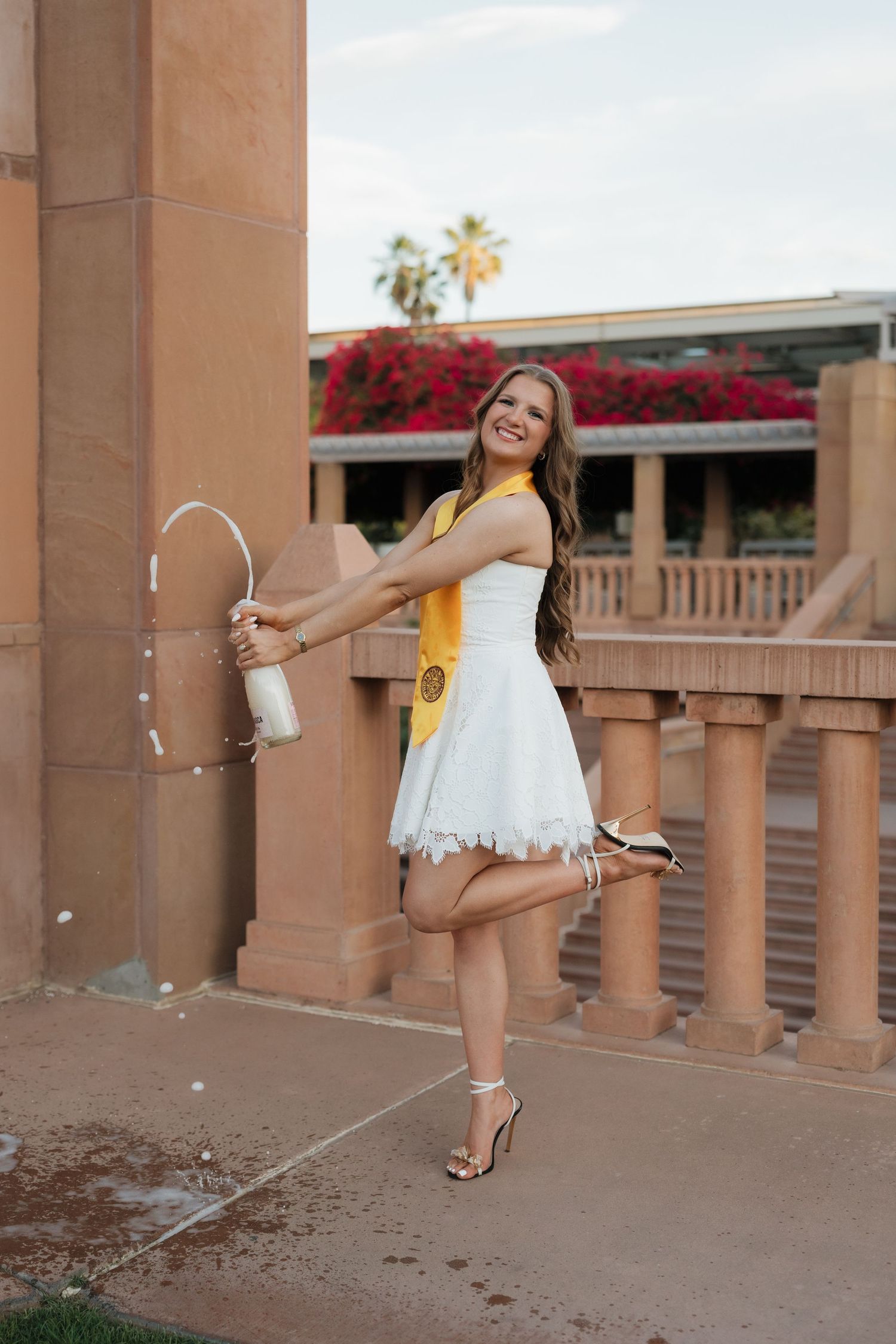Smiling graduate in white dress and yellow sash poses playfully outdoors at a university campus.