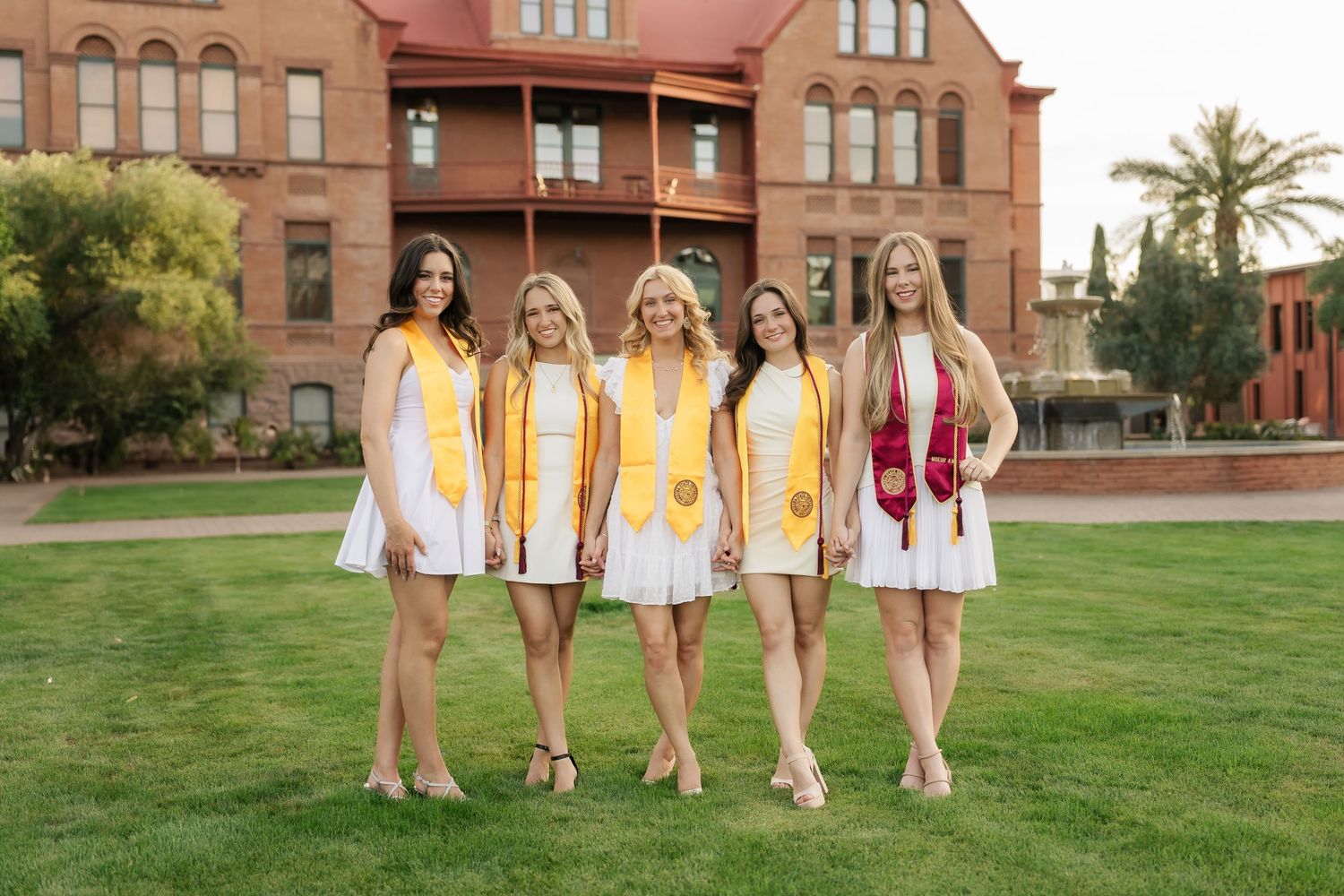 A group poses on grass in coordinated yellow and white dresses for graduation photos.