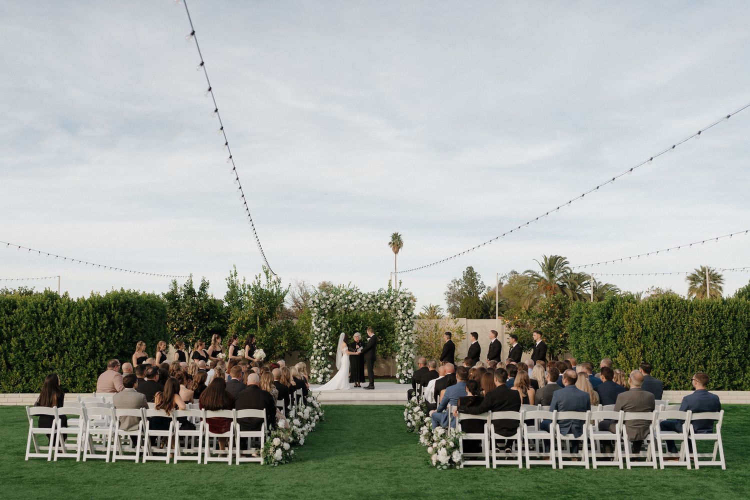 A panoramic view of an outdoor wedding ceremony setup with white chairs and string lights overhead.