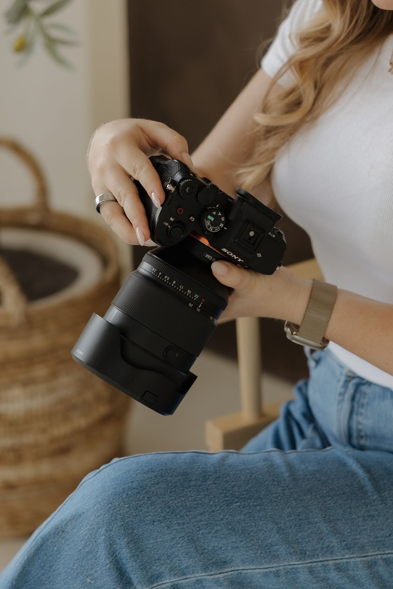 Woman in white top and jeans holding a Sony mirrorless camera with large lens while seated indoors.