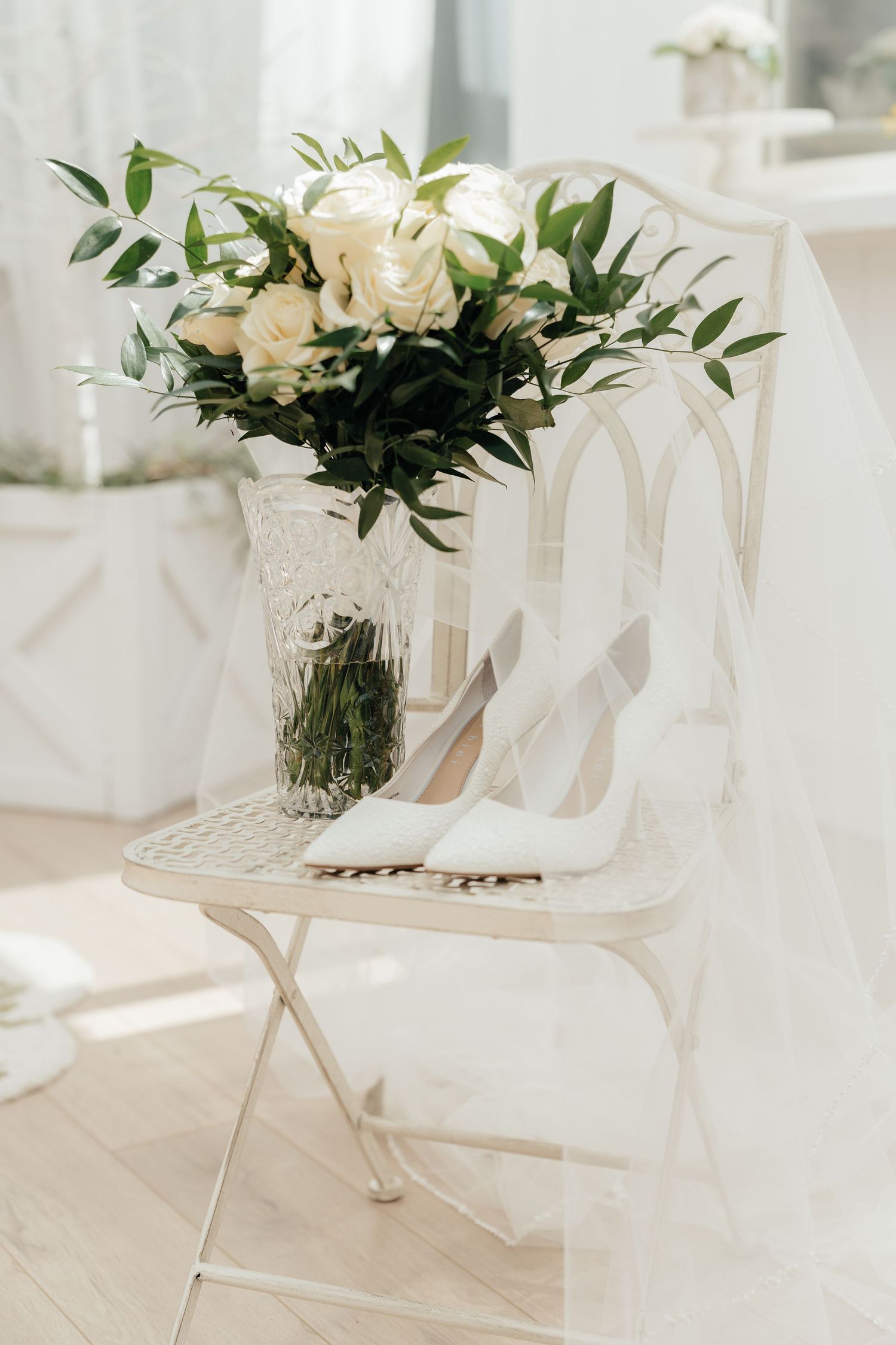 White floral arrangements and elegant bridal shoes displayed on a light wooden floor with soft natural lighting.