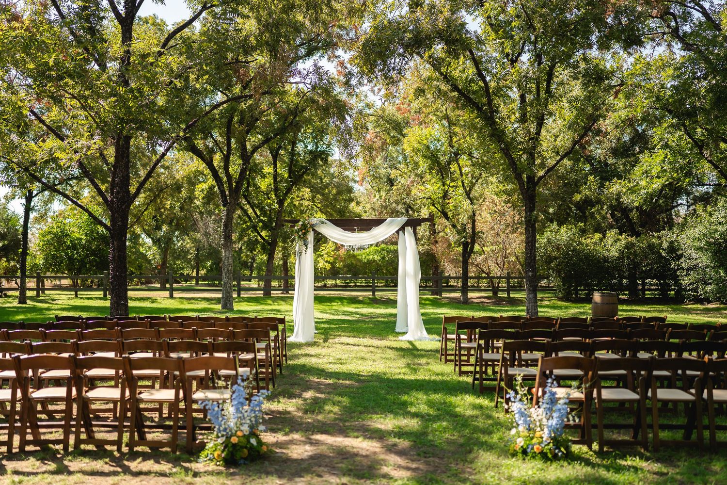 Wooden chairs arranged in rows for an outdoor wedding ceremony under tall trees on a lush green lawn.