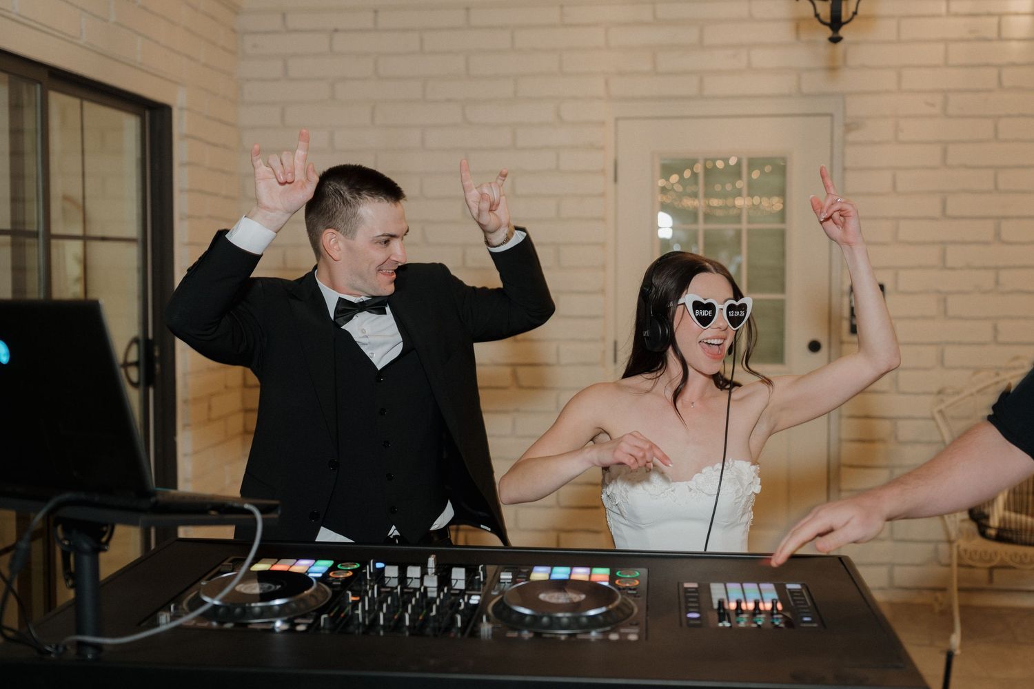 DJ booth at wedding reception with couple dancing and celebrating behind mixing equipment at indoor venue.