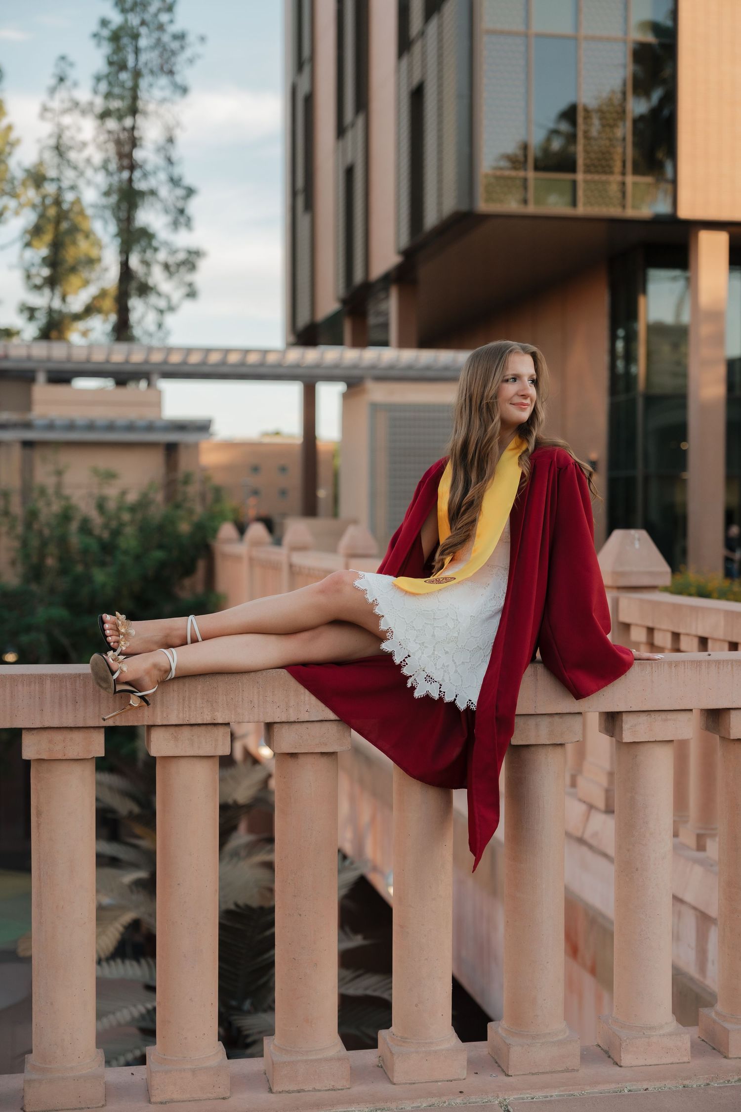 Young woman in graduation gown and yellow sash sits on decorative balustrade outside university building.