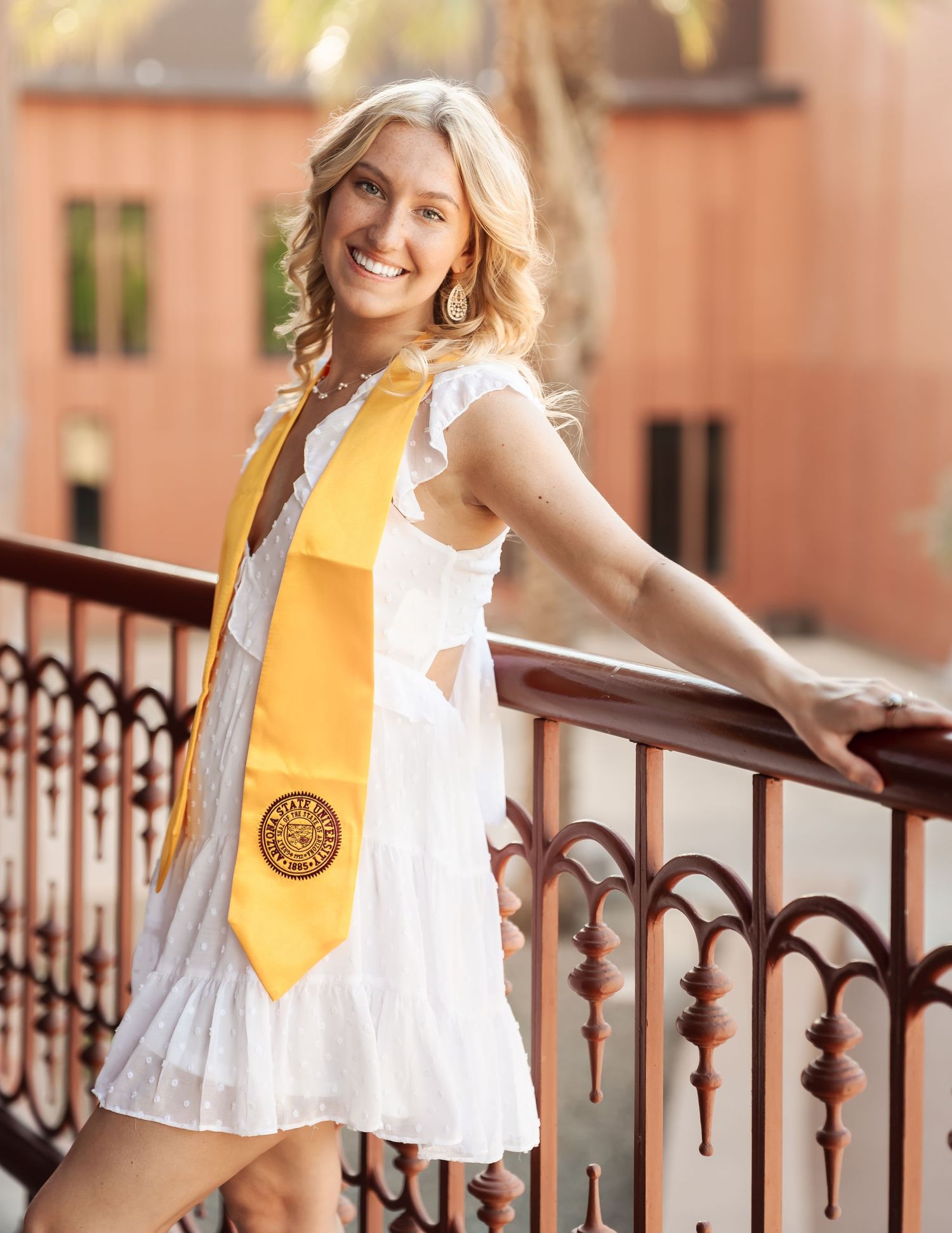 Graduate in white dress and gold stole poses against terracotta building.