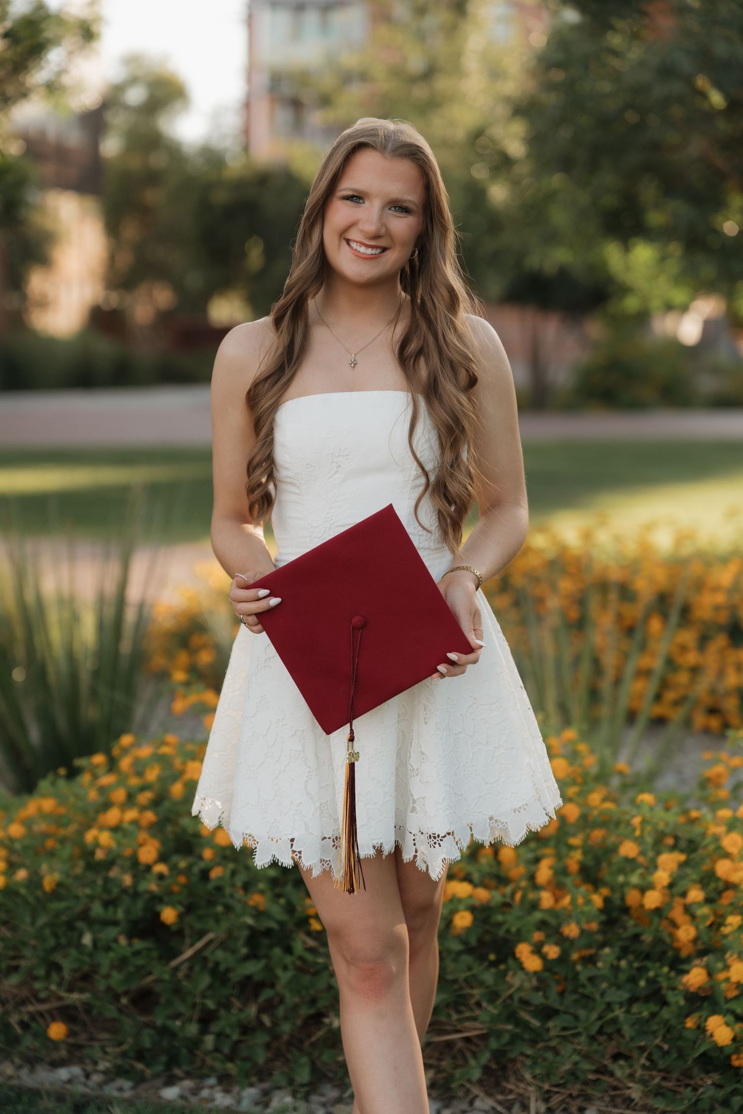 Smiling graduate in white lace dress holding red mortarboard cap outdoors among yellow flowers.