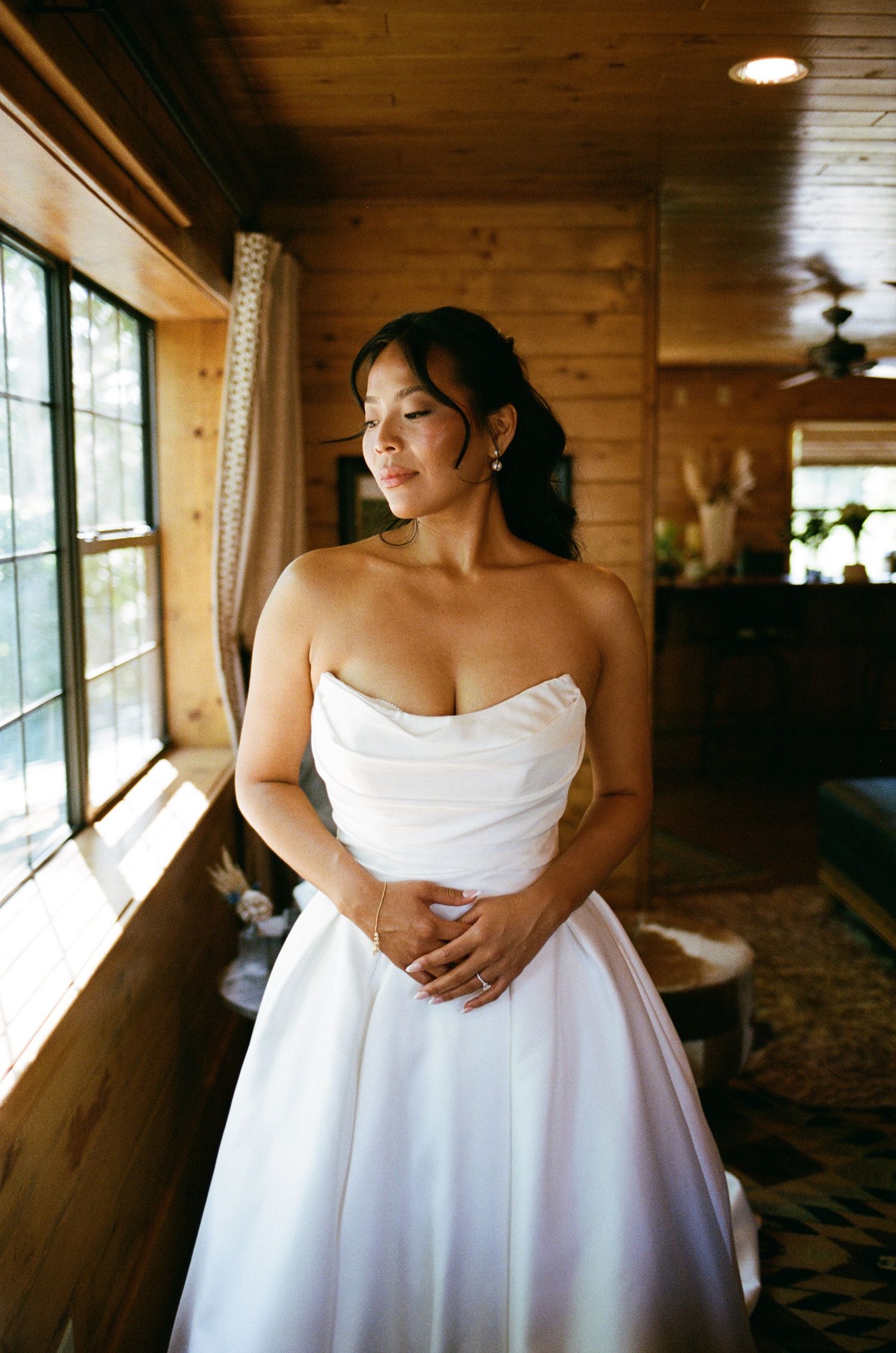 A bride in a strapless white wedding gown gazes out a window in a rustic wood-paneled room with warm sunlight.