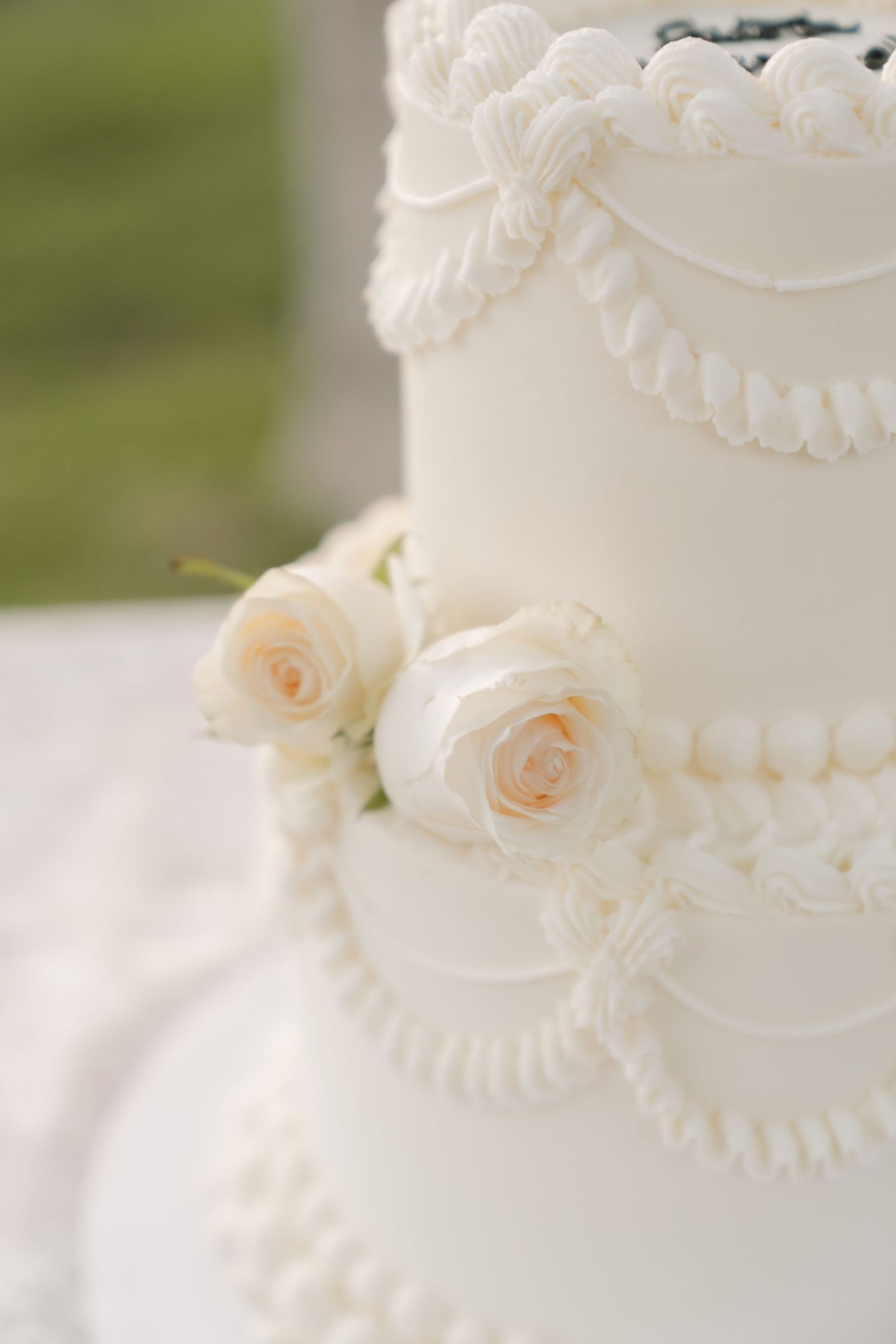 Elegant white wedding cake with cream roses, rope detailing, and pearl borders on a soft green background.