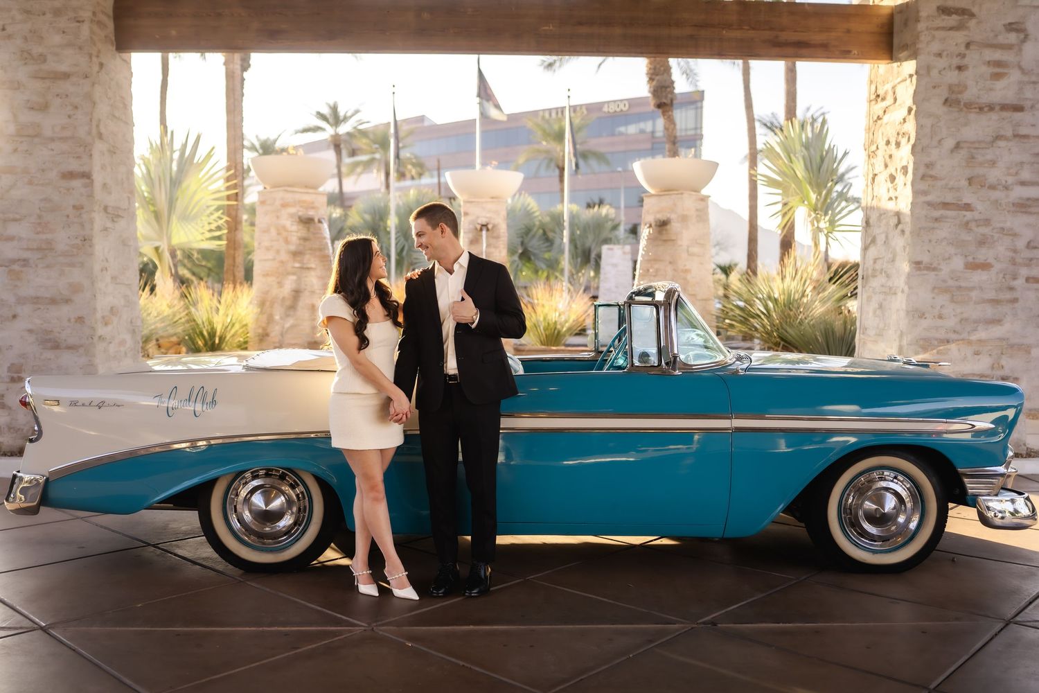 A stylish pair poses beside a classic turquoise convertible car with desert scenery in the background.