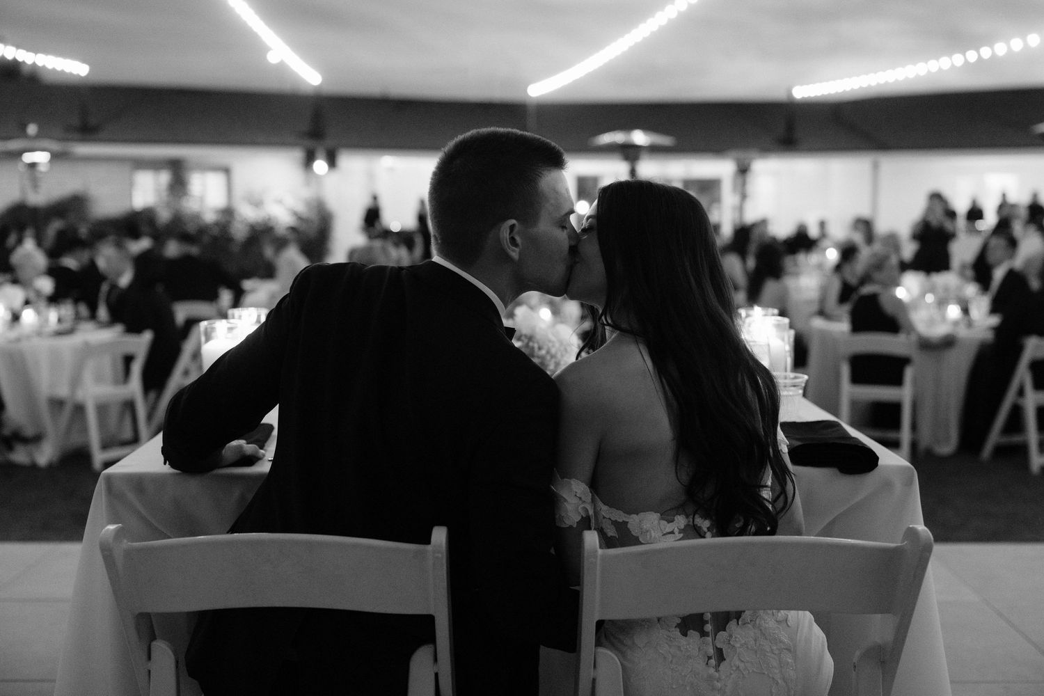Black and white view of wedding guests seated at tables in a large reception venue with overhead lighting.