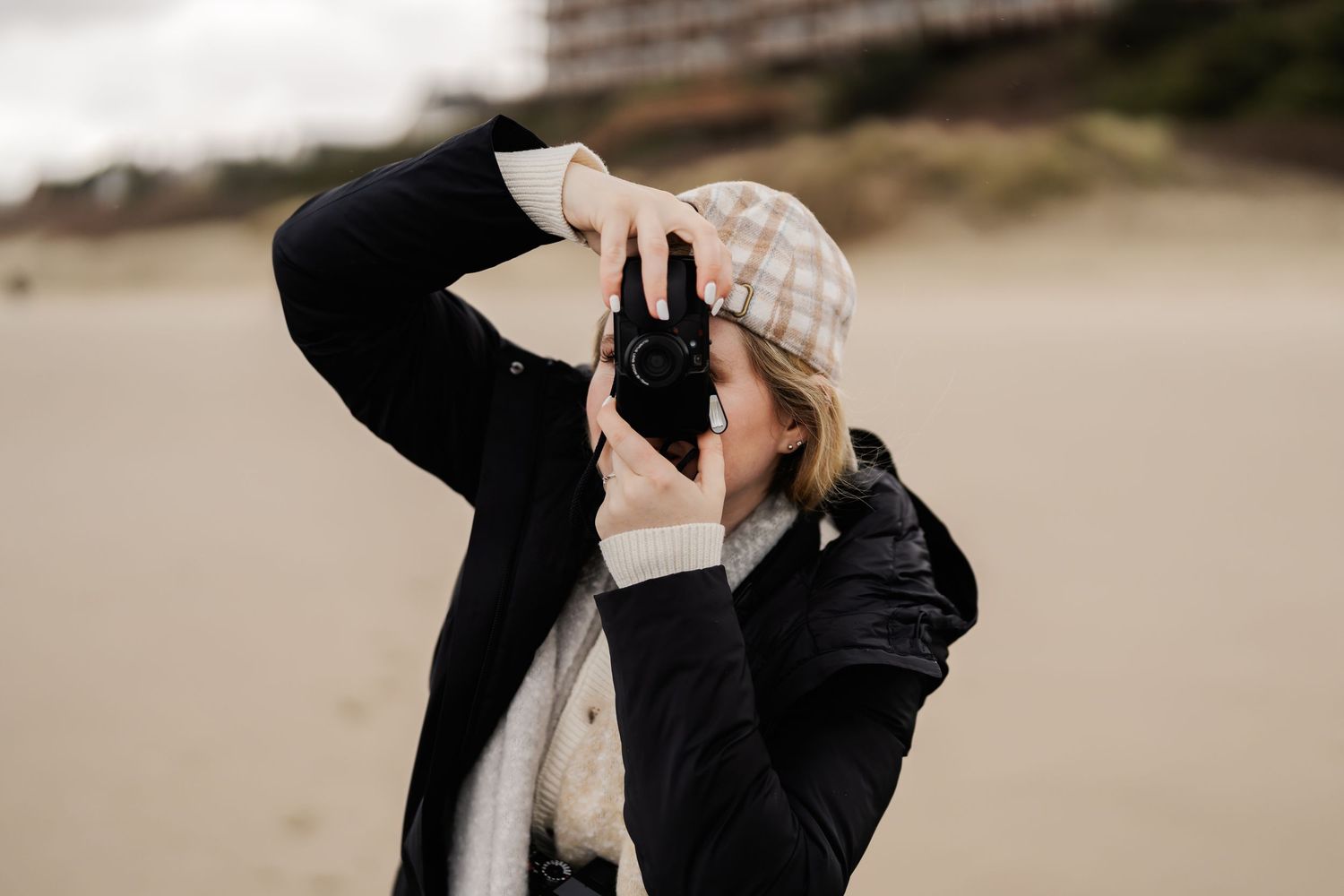 A person in black clothing crouches down to take a photograph outdoors on sandy terrain.