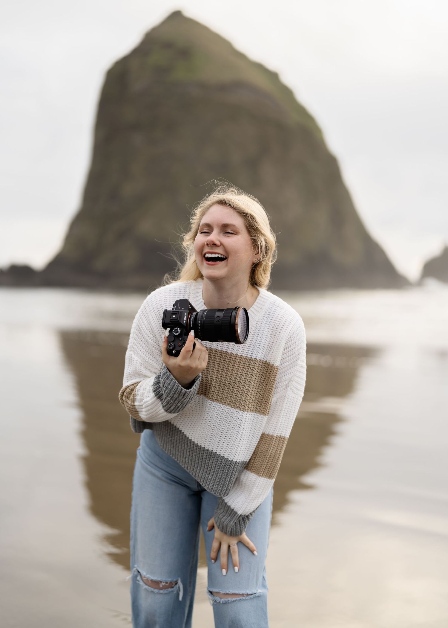 Joyful photographer wearing a striped sweater and ripped jeans laughs while holding camera at Cannon Beach, Oregon.