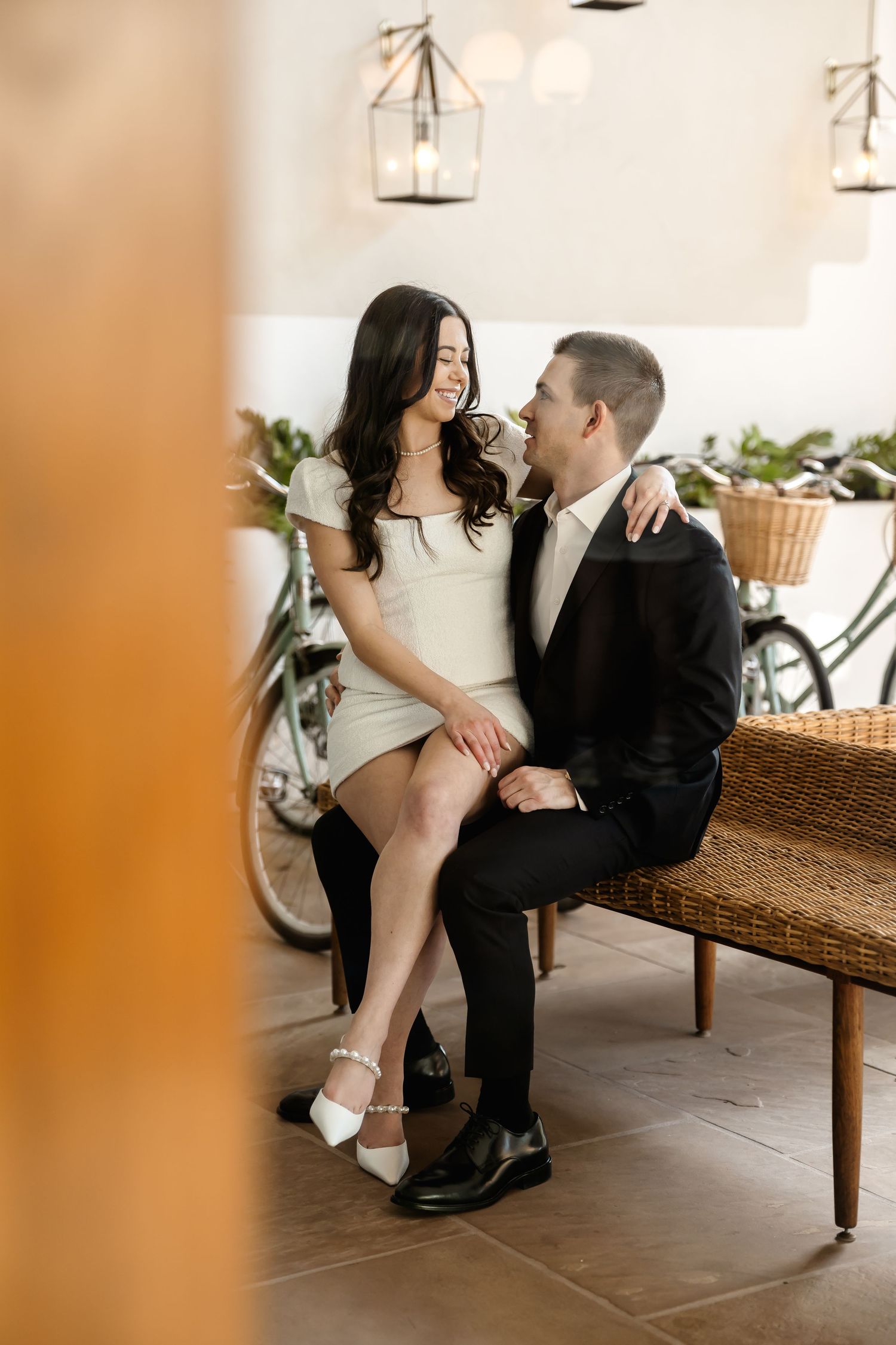 Couple sharing tender moment on bench with decorative lighting and plants in background.