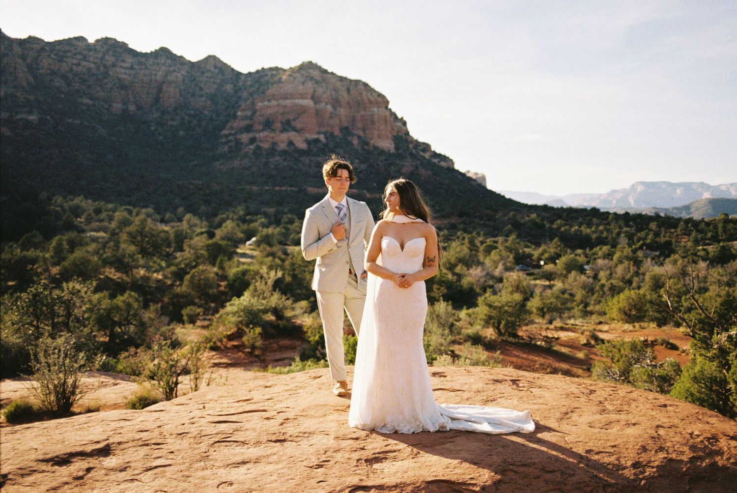 Couple in wedding attire standing on red rock in Sedona, Arizona, with desert landscape and red mountains behind them.