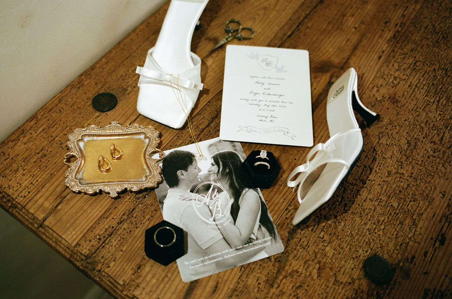 Wedding flat lay with white heels, rings, invitation, and romantic black-and-white couple photo on rustic wood table.