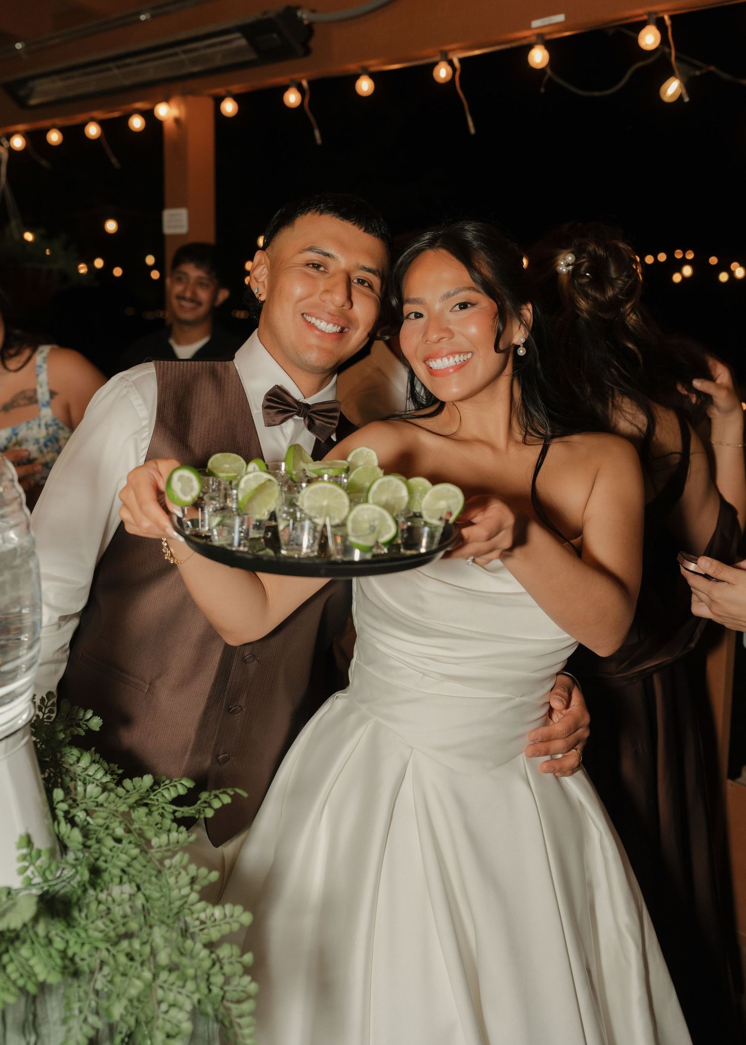 Newlyweds smiling and holding a tray of tequila shots with lime at their wedding reception.