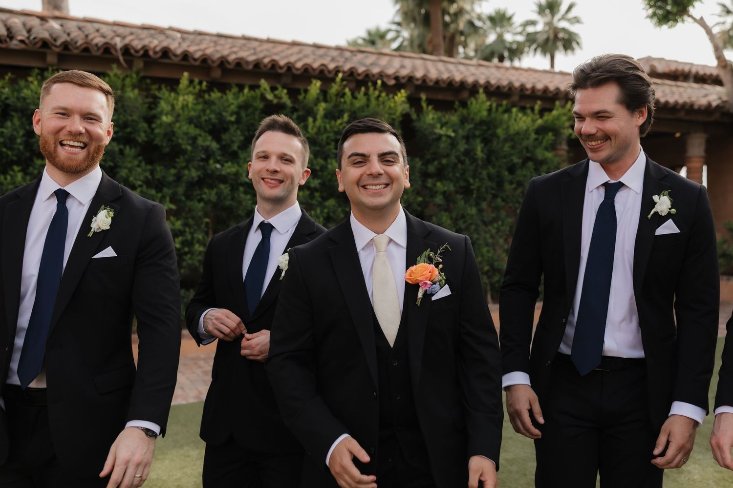 Groom in beige suit with floral boutonniere poses with three groomsmen in black suits outdoors at wedding.