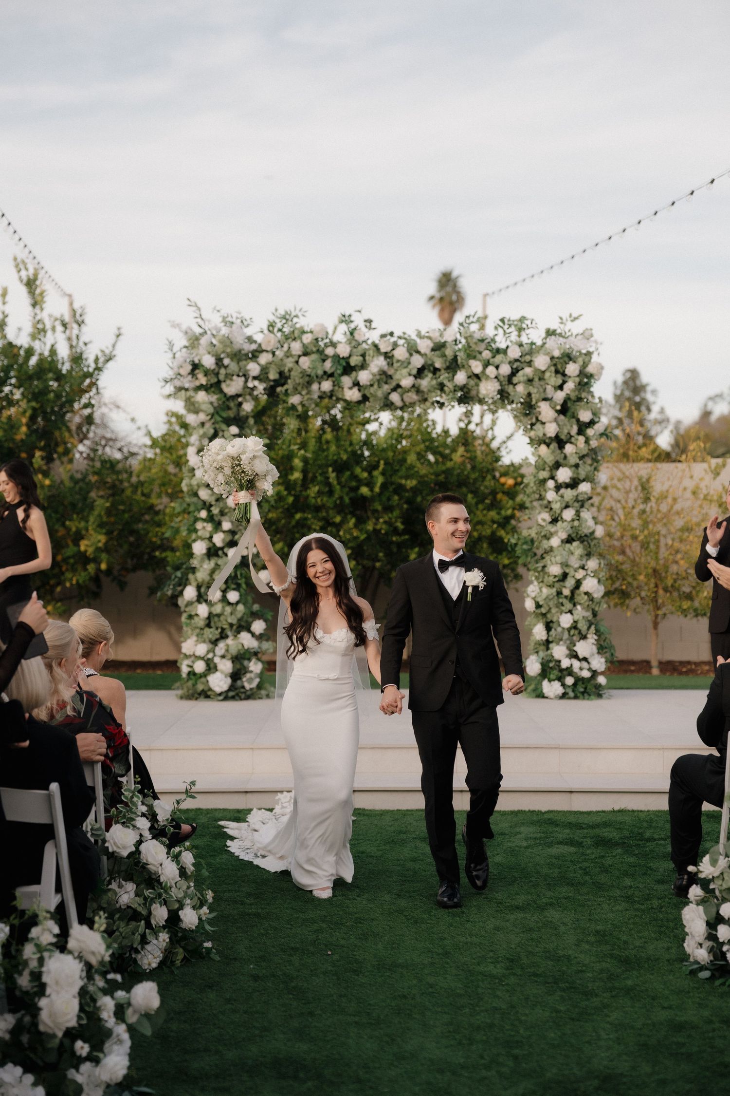 Wedding ceremony exit under floral arch with white roses as guests celebrate on green lawn.