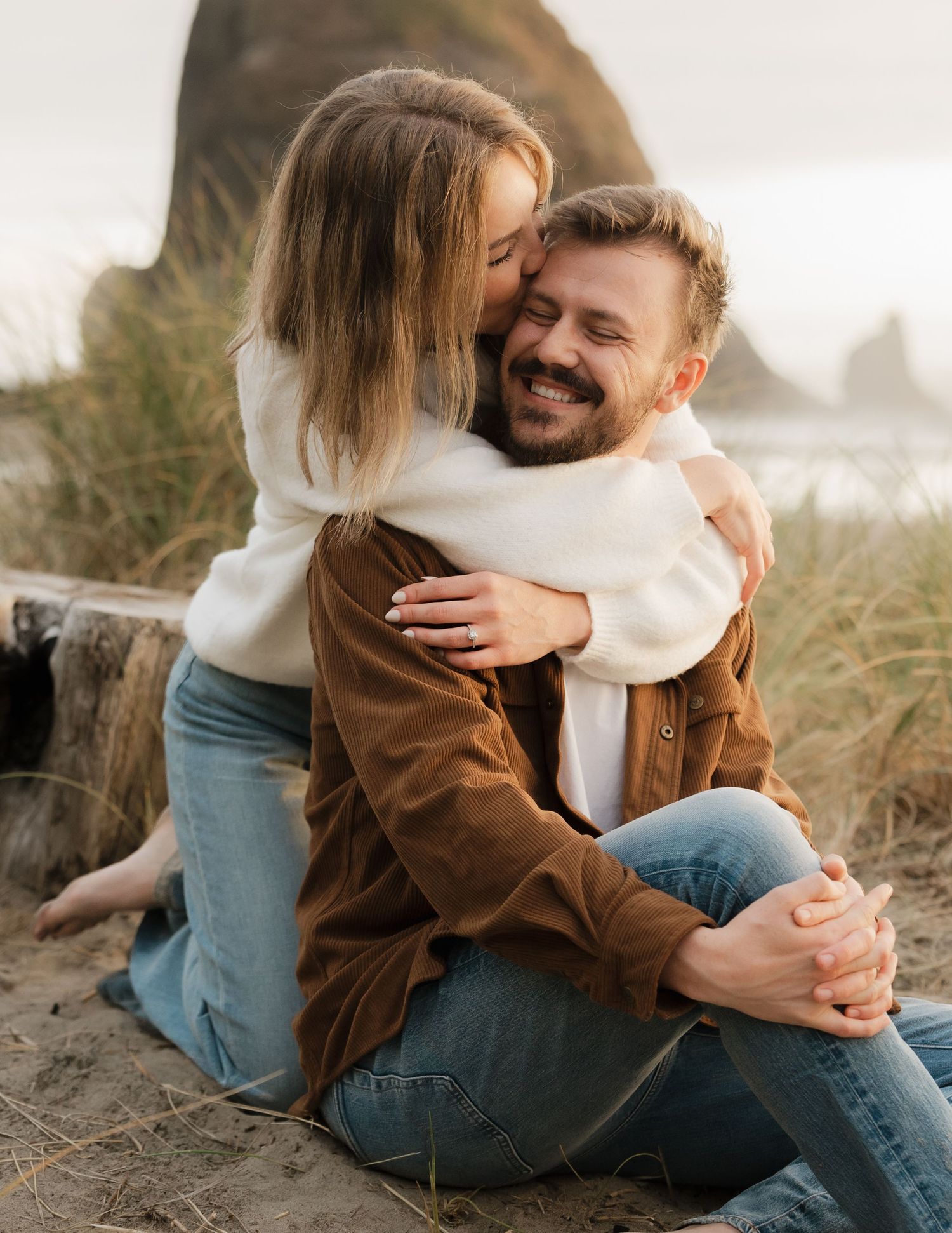 A happy couple embraces on a sandy beach, woman hugging man from behind, both smiling, with rocky coastline in background.